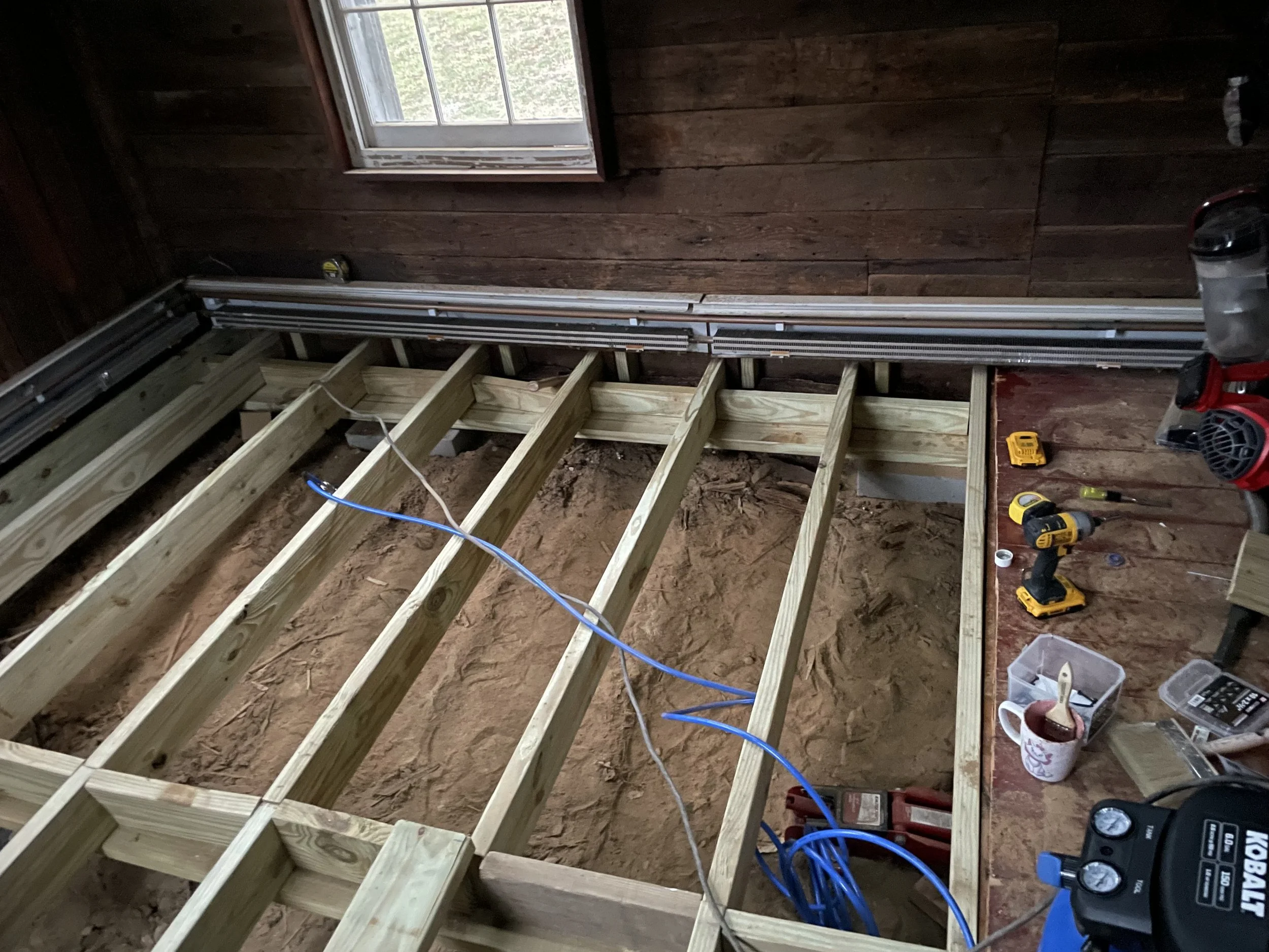 Interior of a room under construction with wooden framing, a window, and electrical wiring, tools, and construction materials scattered on the floor.