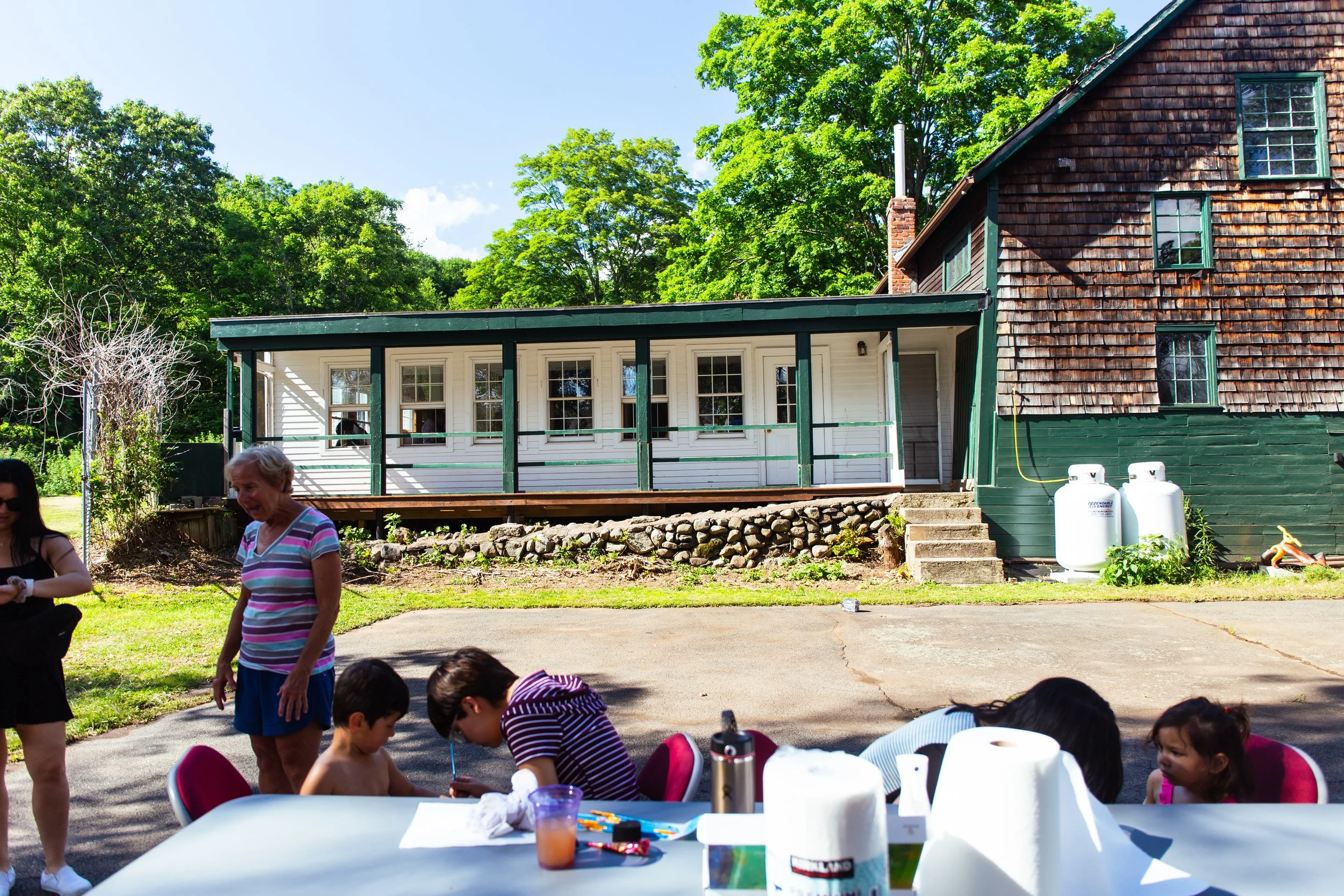 Children and adults gathered outdoors near a table with paper towels and art supplies, with a house and green trees in the background.