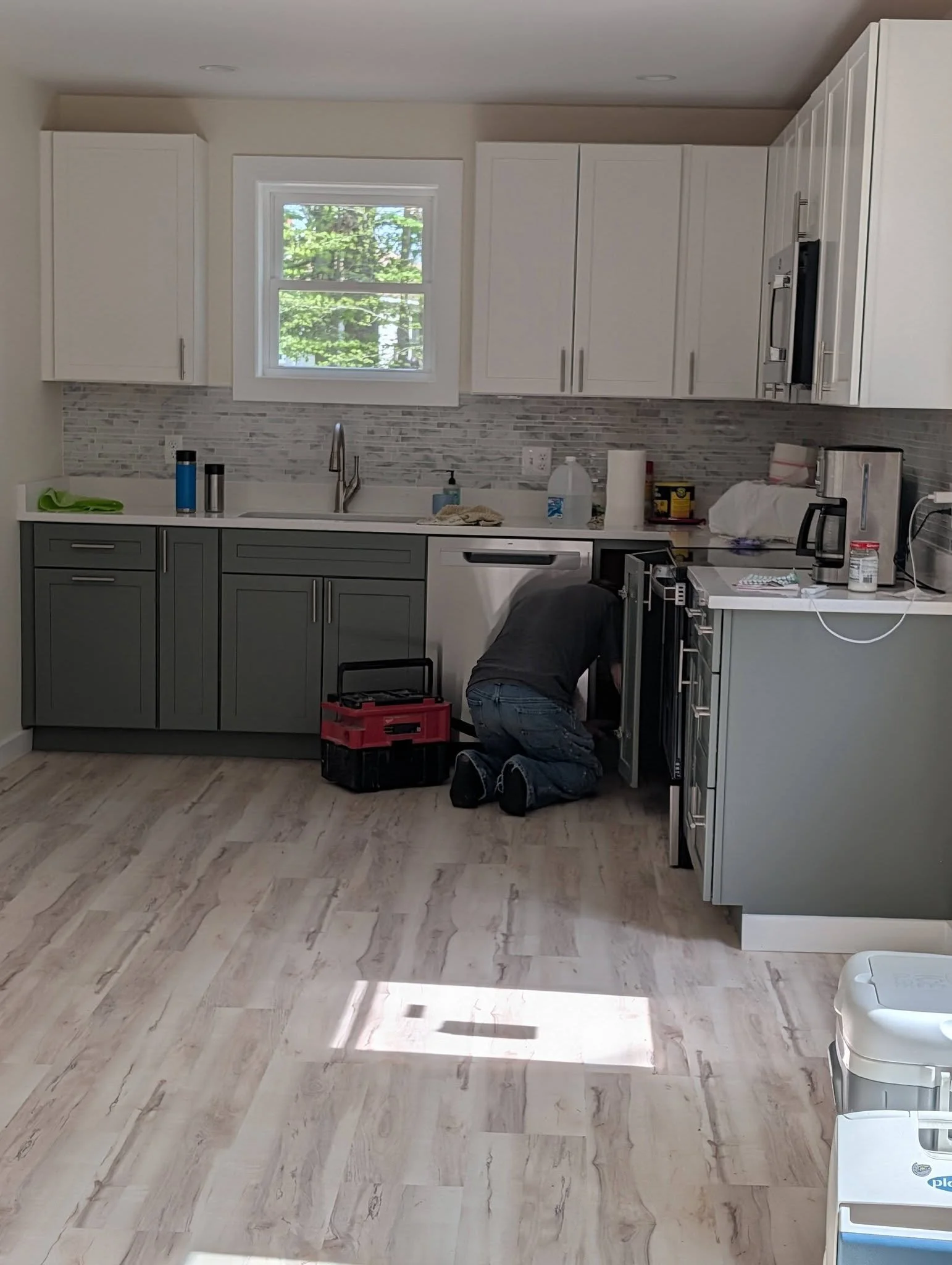 Kitchen with gray and white cabinets, a window with trees outside, and a person working under the sink.