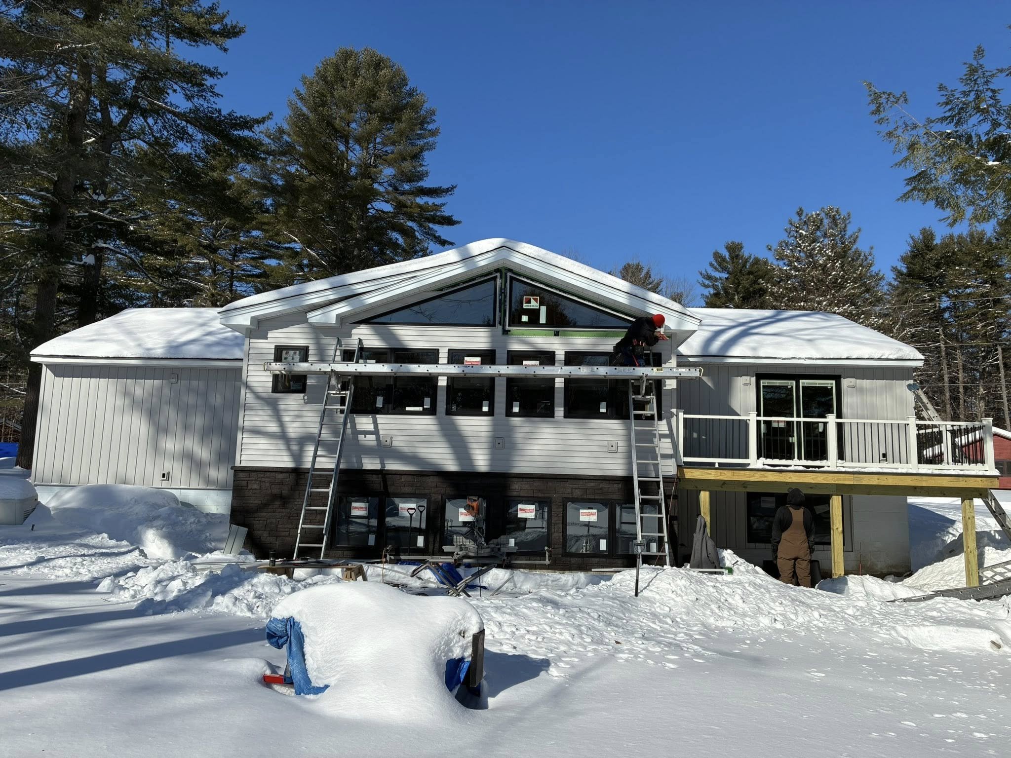 Construction workers building a house in a snowy landscape, with tall pine trees and a clear blue sky in the background.