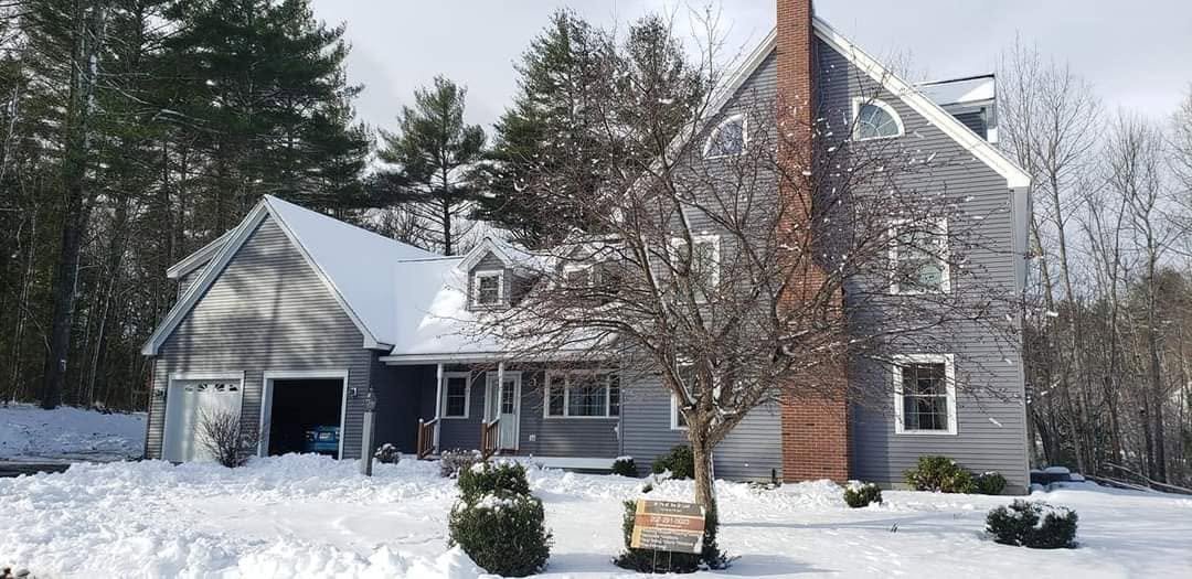 A large house with gray siding and a brick chimney in a snowy landscape, with a tree and bushes in front.