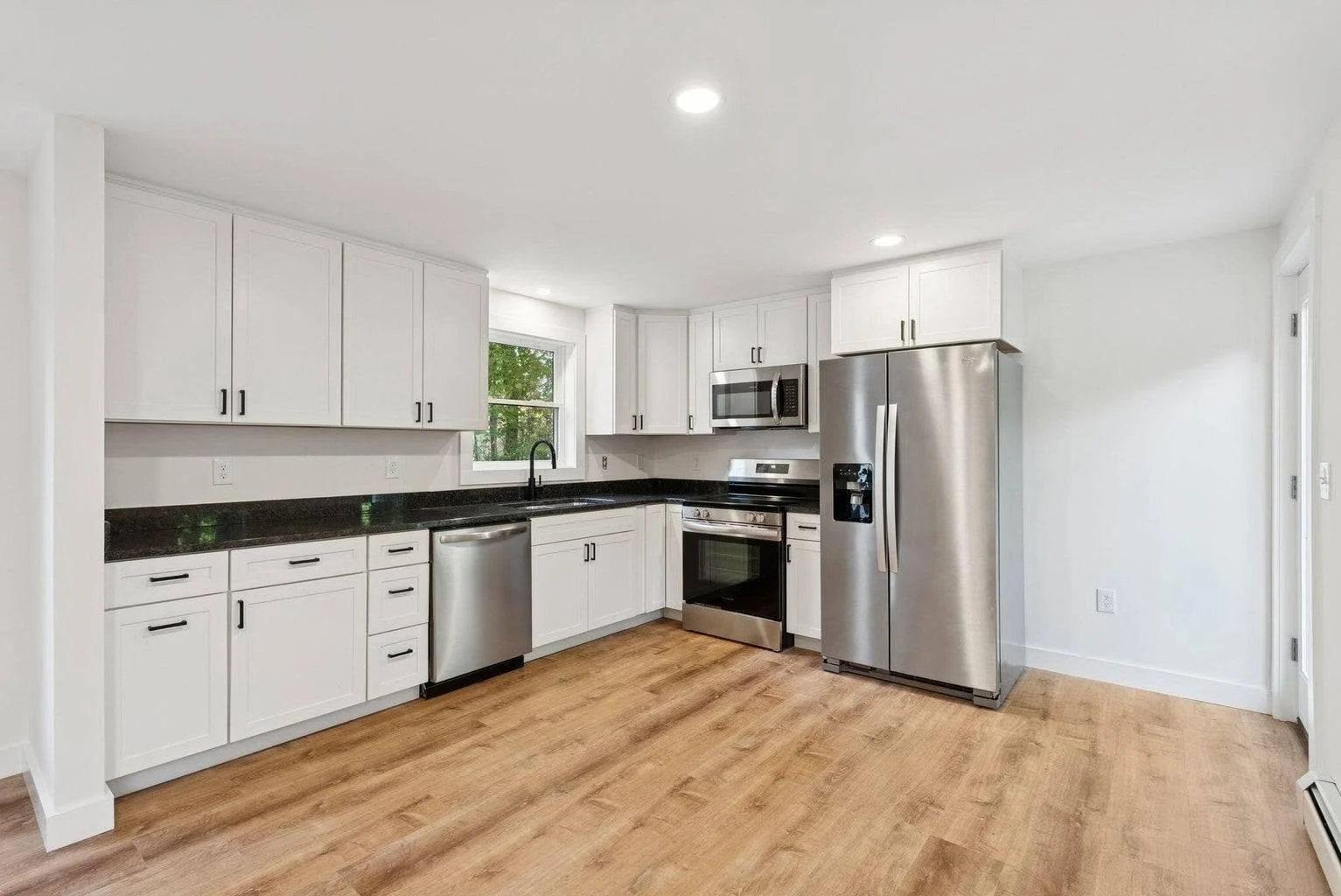 Modern kitchen with white cabinets, black countertops, stainless steel appliances including a refrigerator, microwave, oven, and dishwasher, and wood flooring.