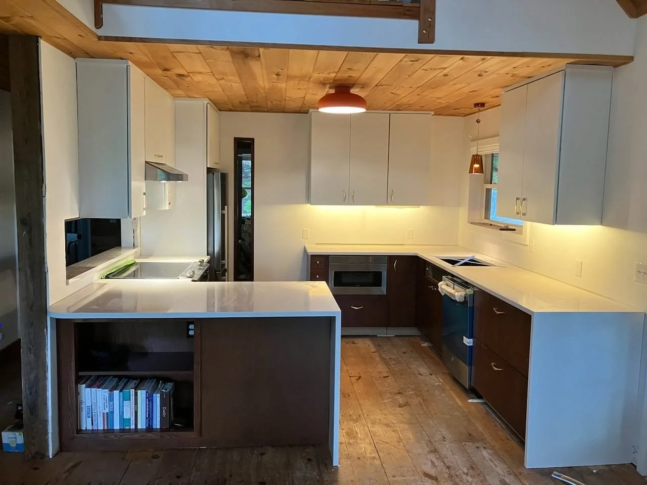 Modern kitchen with white and dark brown cabinets, white countertops, wooden ceiling, and wooden floor. Features built-in appliances, small island with books, a window, and contemporary lighting fixtures.