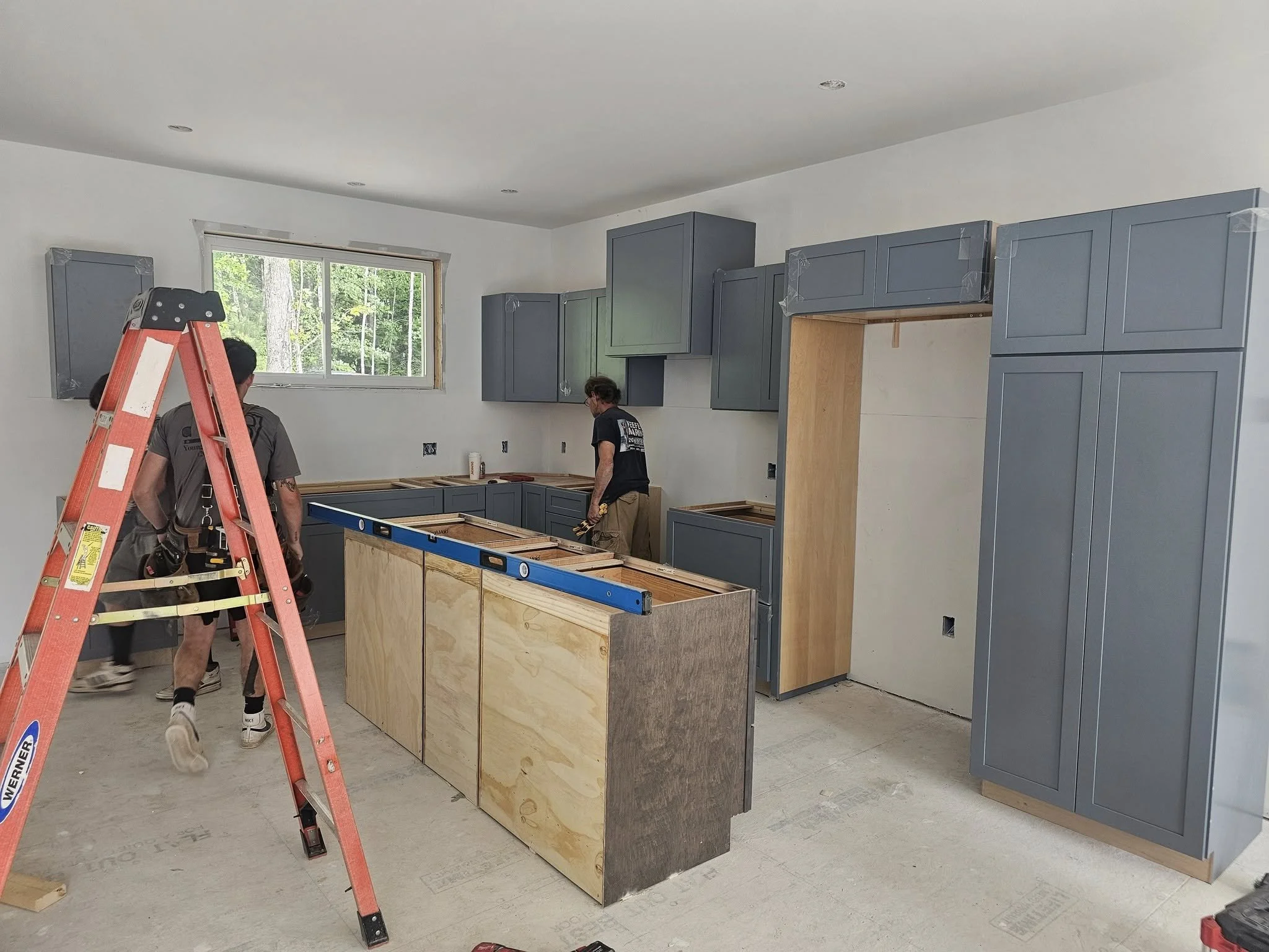 Kitchen under renovation with partially installed blue cabinets, a plywood island frame, a ladder, and two workers.