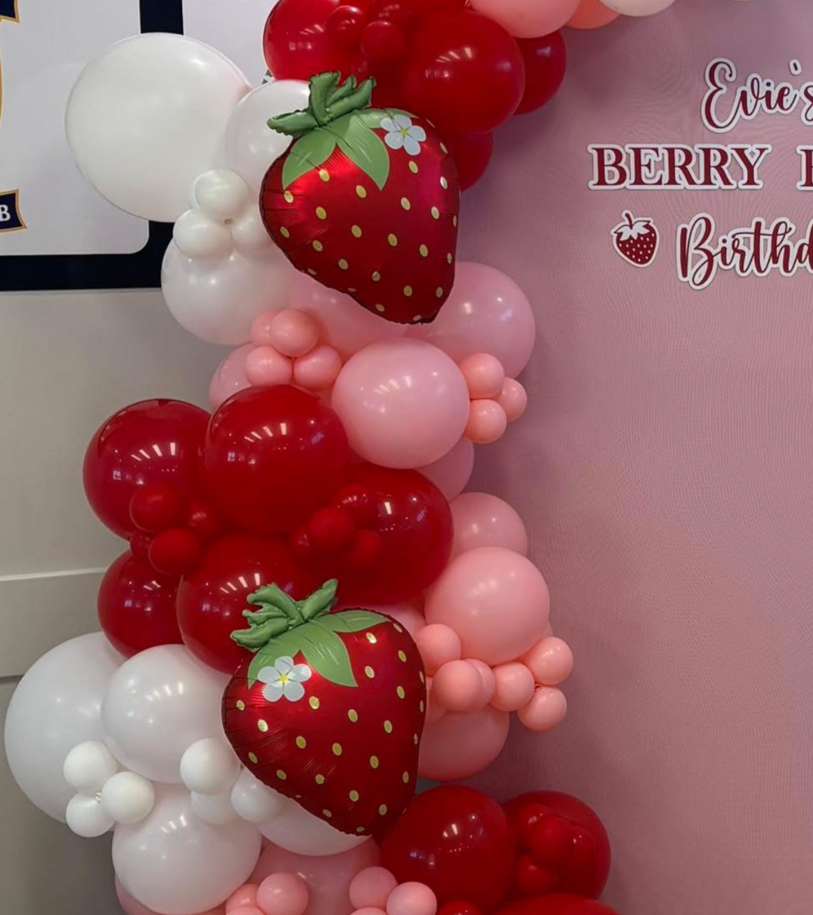 Balloon decoration featuring red, white, and pink balloons with two large strawberry-shaped balloons, and a pink backdrop with text that reads 'Berry Birthday'.
