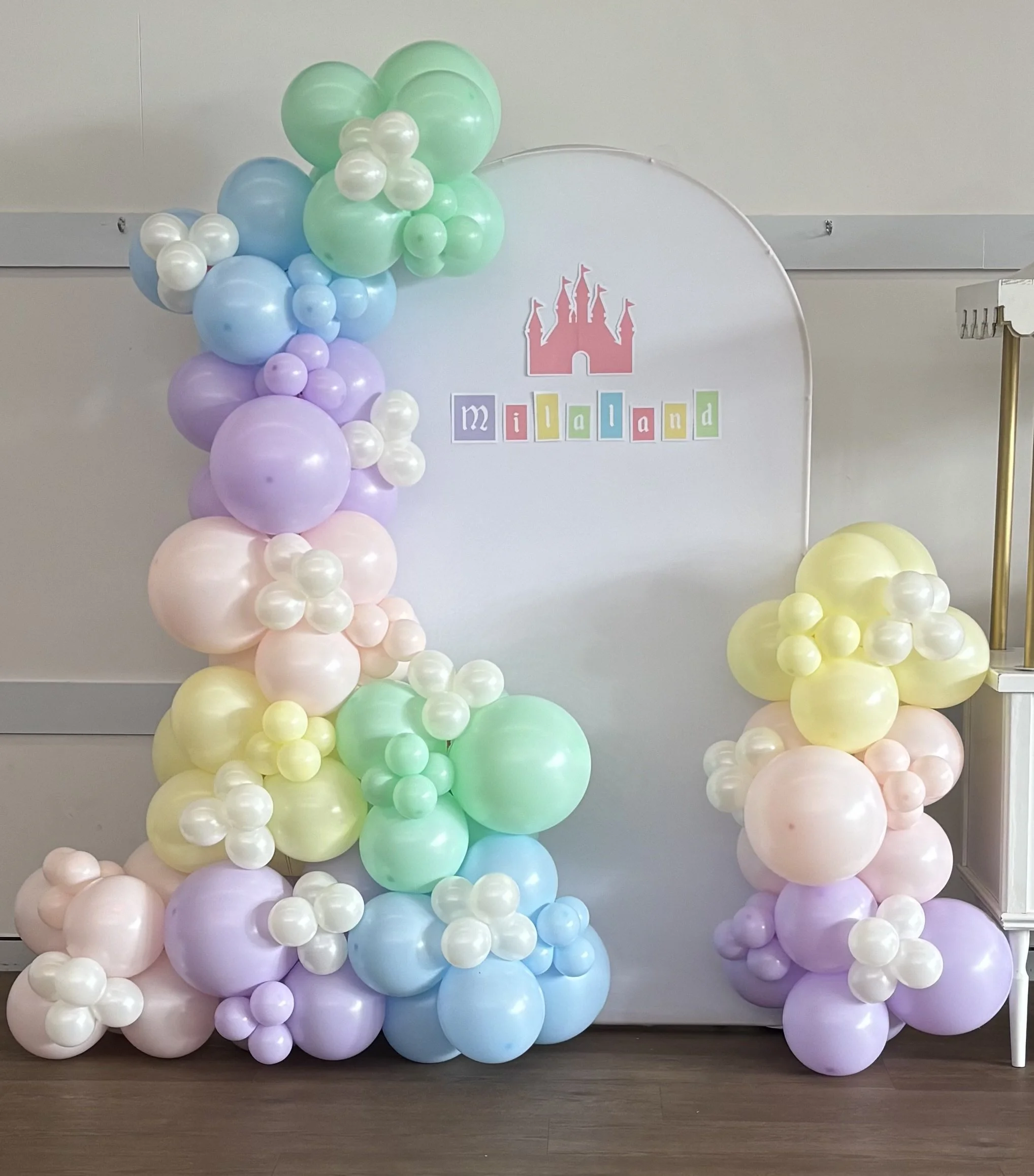 Decorative balloon arrangement in pastel colors including purple, yellow, green, blue, and white, arranged in clusters and featuring a pink castle cutout with colorful blocks spelling 'mialand' in the background.