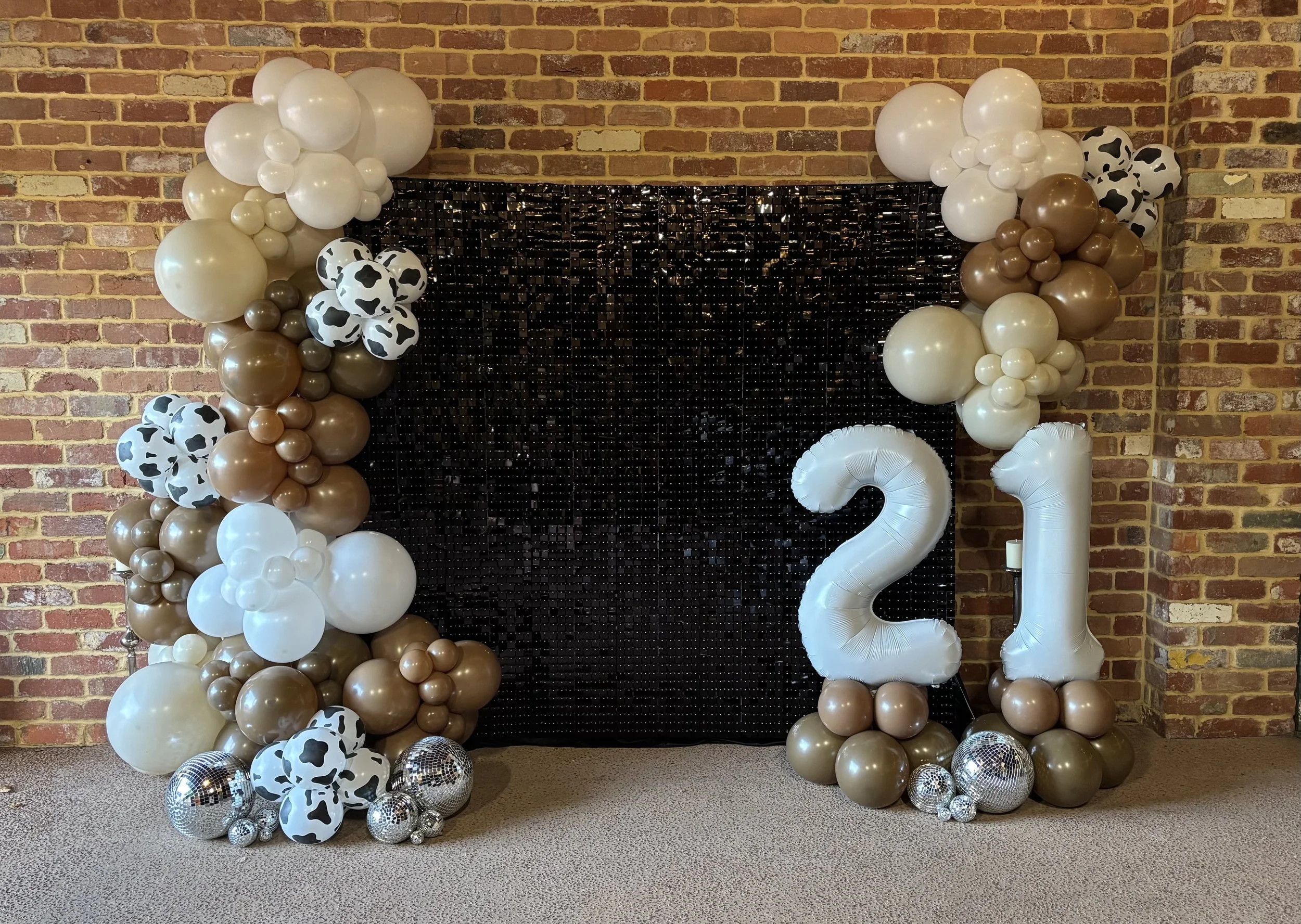 Balloon decoration with cowskin pattern balloons, gold, silver, white balloons, and are a part of the backdrop for a celebration, with a black sequin backdrop and a '2023' balloon, set against an exposed brick wall.