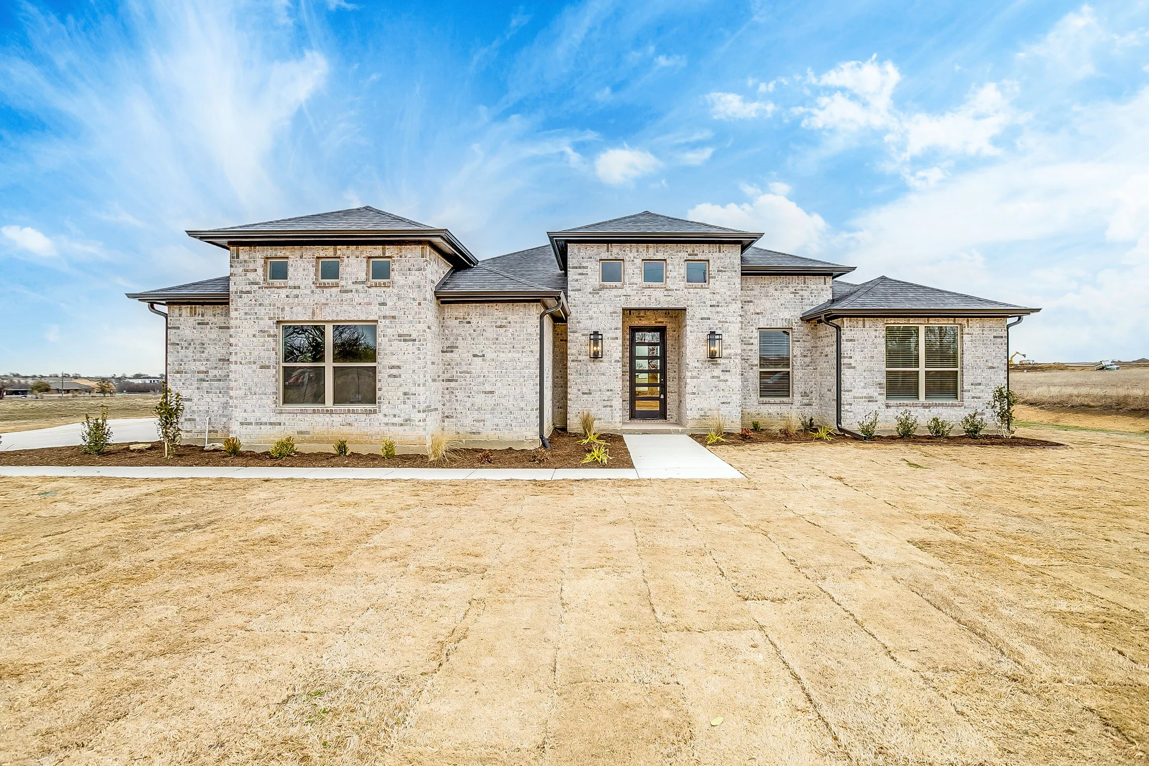 Newly built brick house with a modern design, large windows, and a black front door, set on a landscape with patchy grass and a clear blue sky.