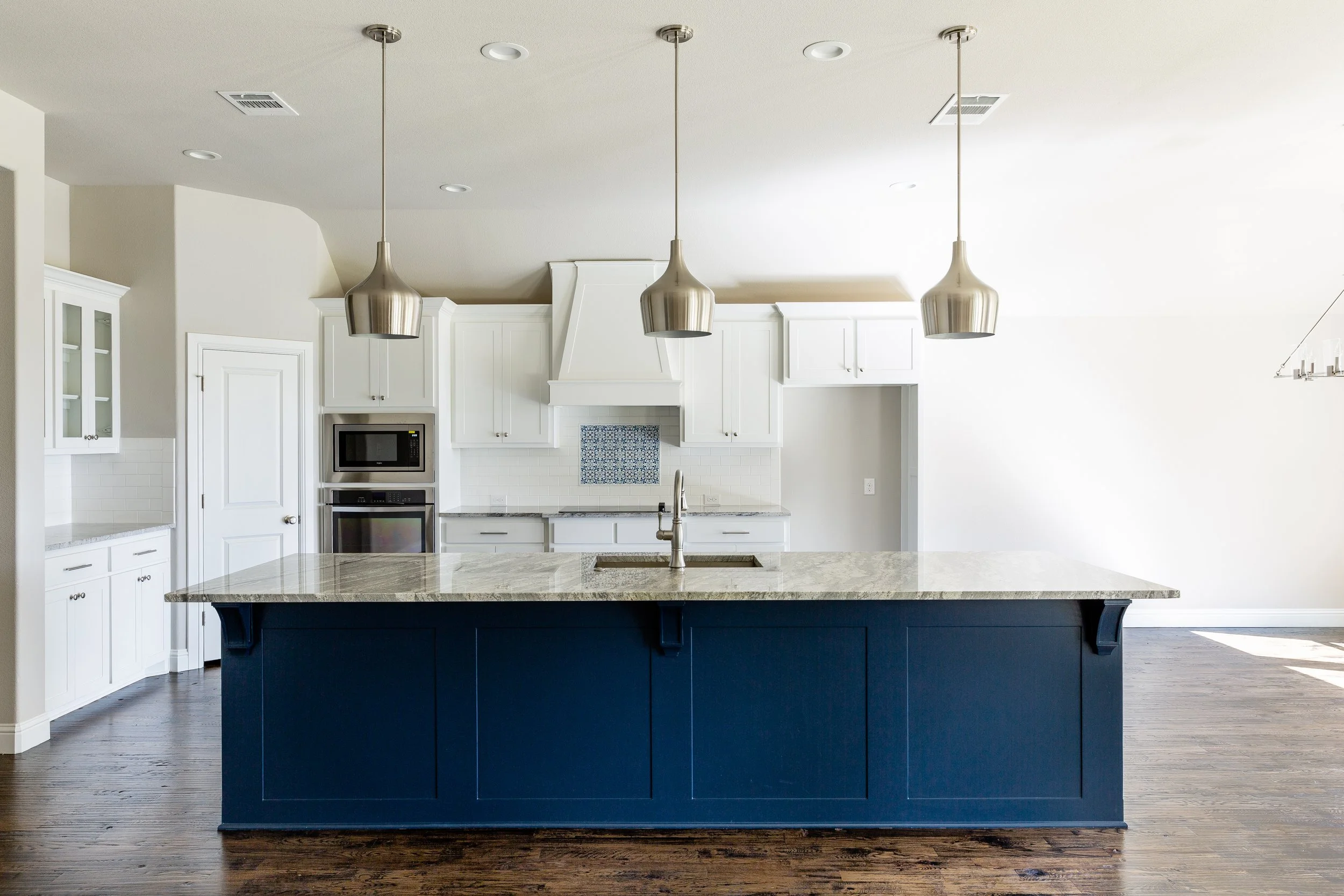 Modern kitchen with white cabinets, stainless steel appliances, and a dark blue island with a light granite countertop, pendant lights, and hardwood flooring.