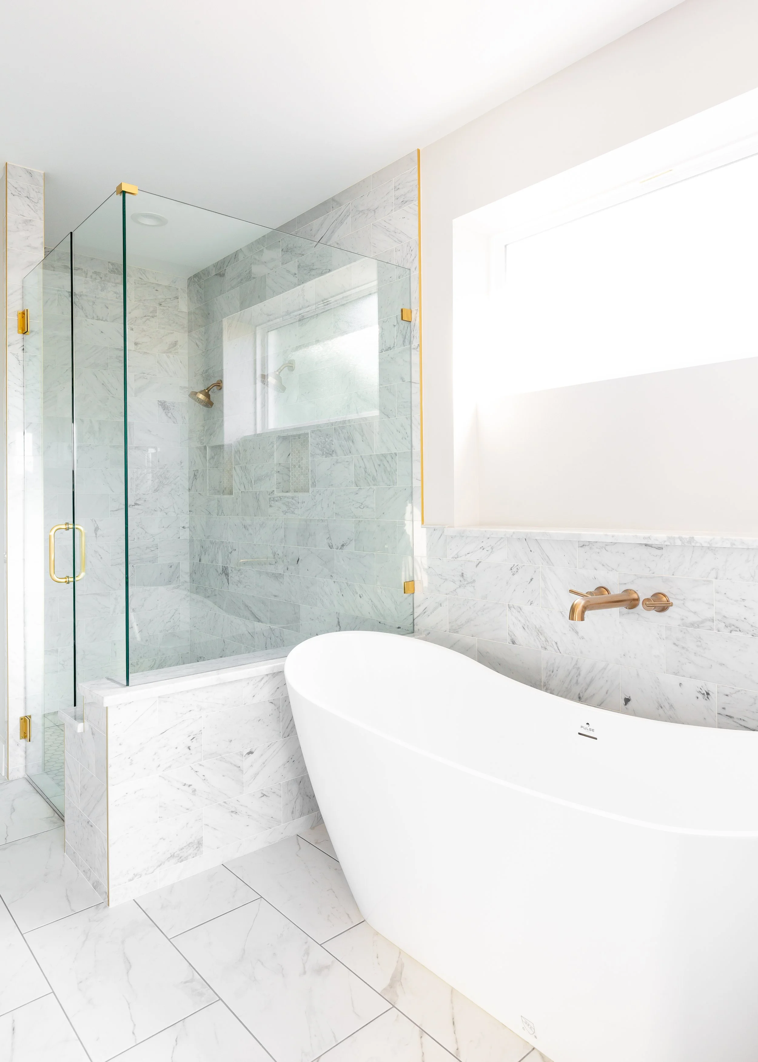 Modern bathroom with a white freestanding bathtub, marble walls and floors, and a glass-enclosed shower with brass fixtures and a window.