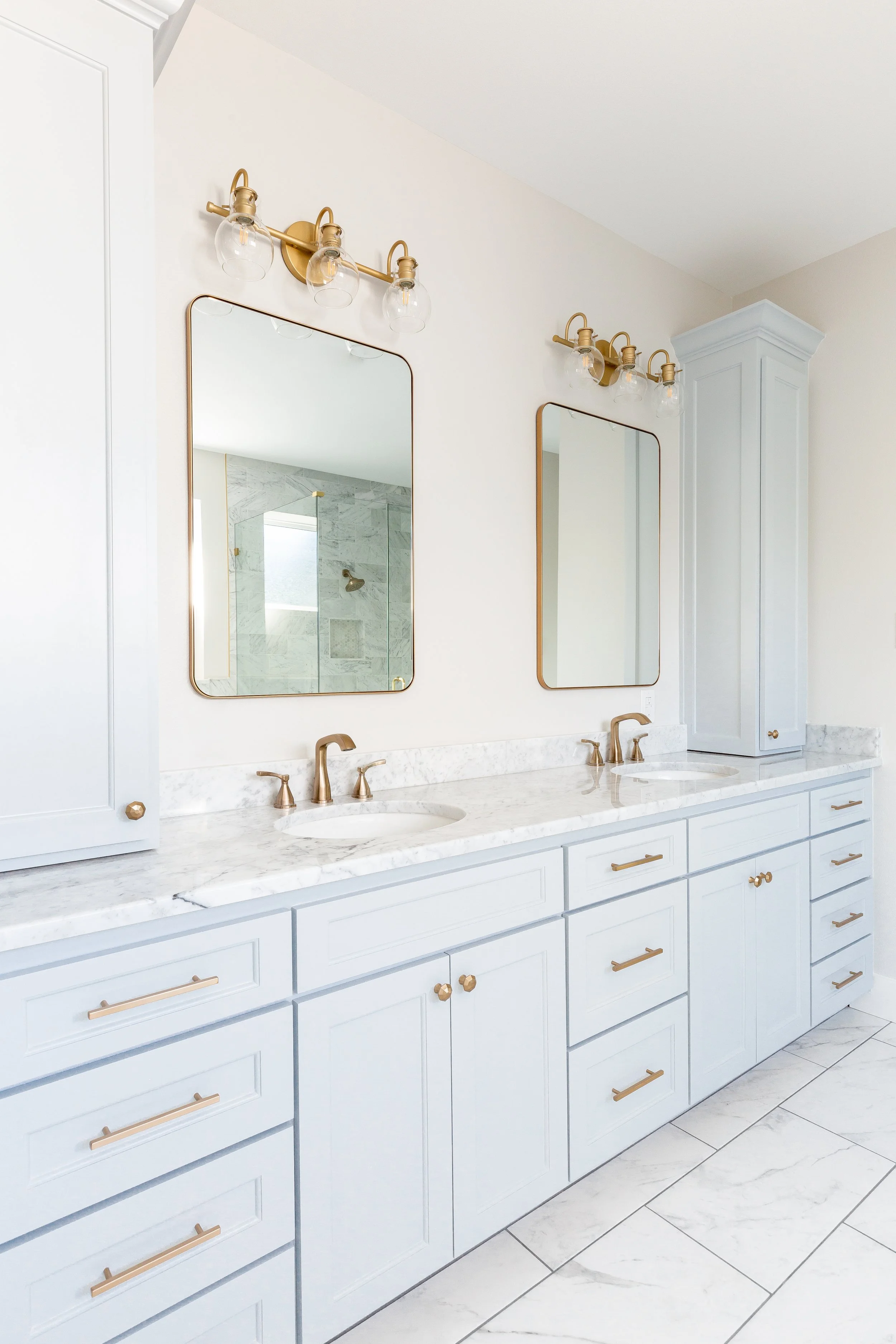 Clean white bathroom with double vanity, marble countertop, gold fixtures, two rectangular mirrors, and a shower visible in the mirror reflection.