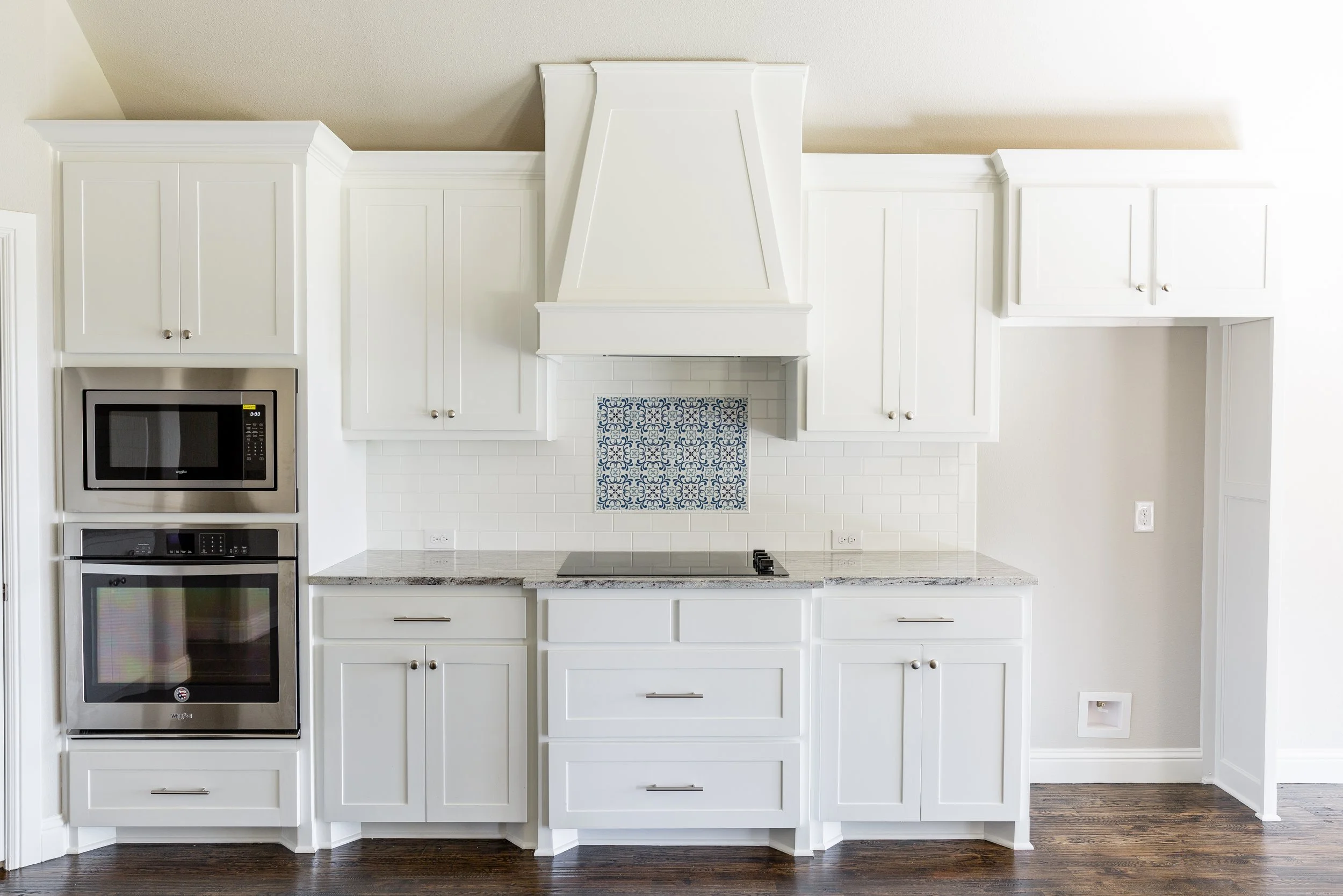 White kitchen cabinets with a built-in microwave and oven on the left, a white range hood in the center, and a backslash with a blue and white pattern behind the stove, all under a beige ceiling and dark hardwood floor.