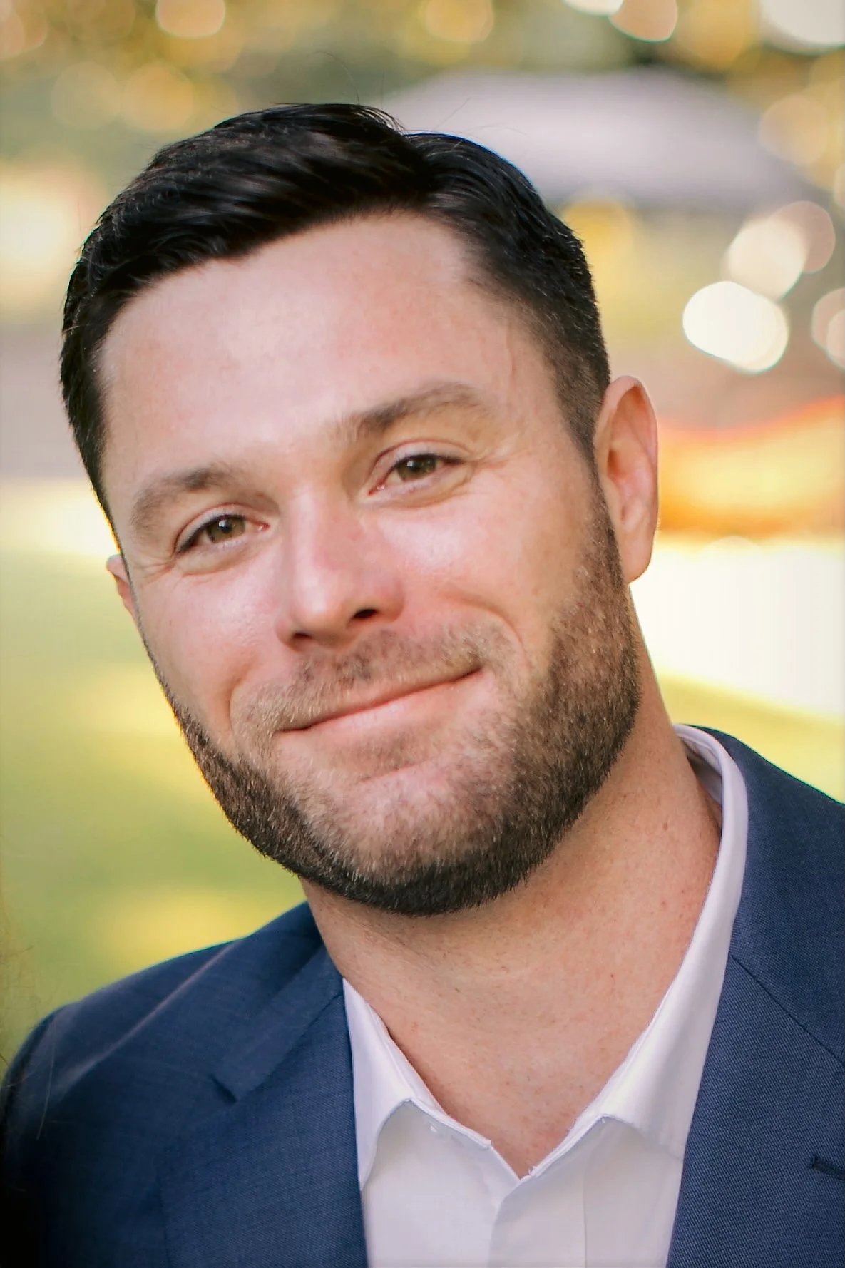A man with short dark hair, a beard, and light skin smiling outdoors with blurred lights in the background, wearing a dark blue suit jacket and a white dress shirt.