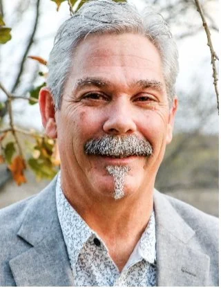 A smiling older man with gray hair and a mustache, wearing a gray suit and patterned shirt, standing outdoors with trees and a fence in the background.