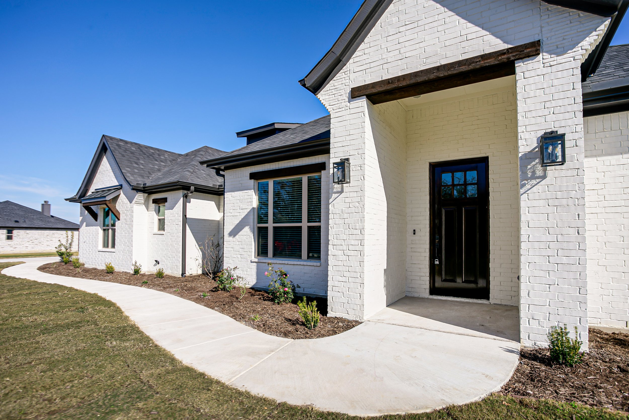 Front view of a modern white brick house with black front door, black window frames, and exterior wall-mounted lantern lights. Curved concrete pathway leads to the entrance, surrounded by small landscaped plants and lawn, under a clear blue sky.