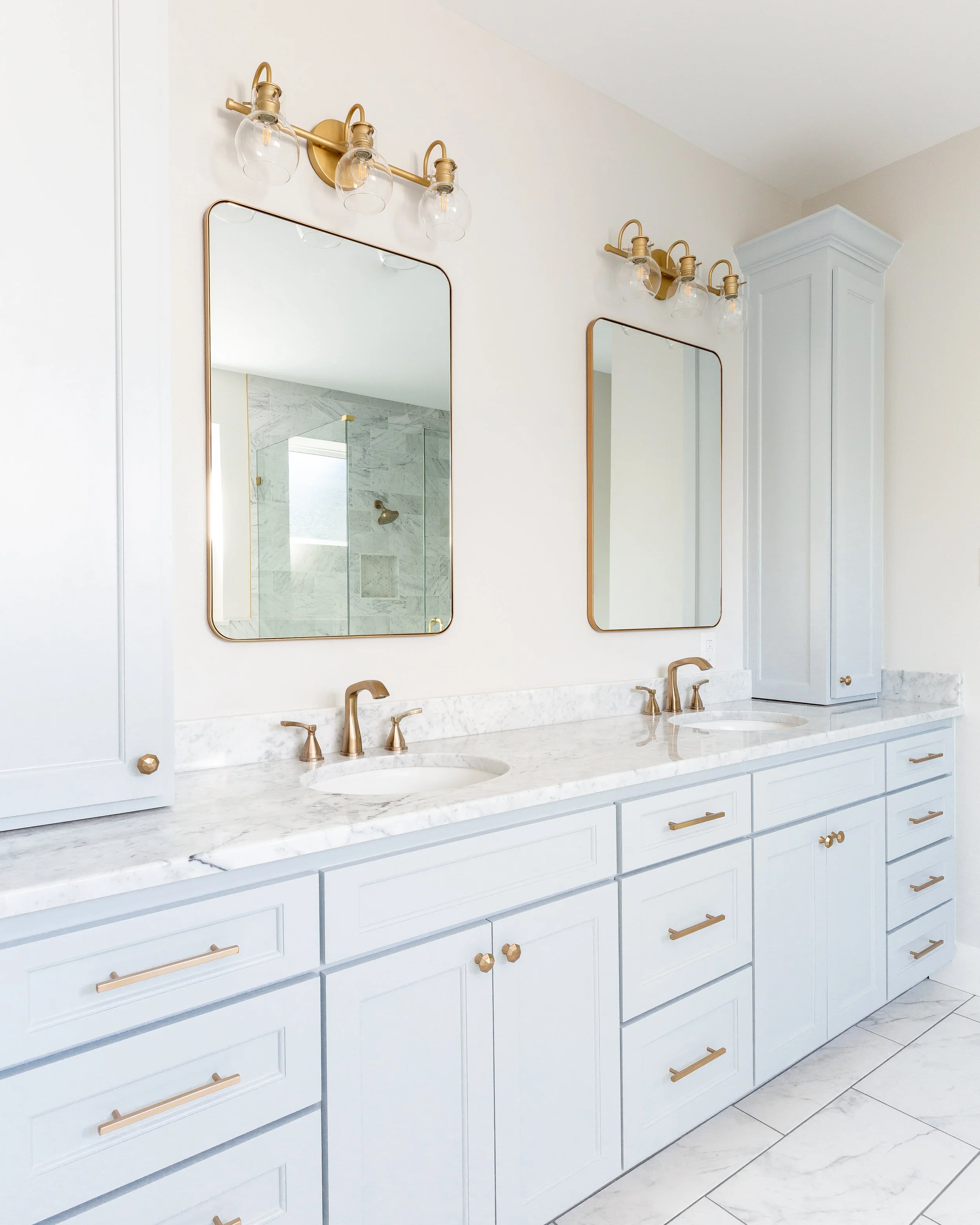 A bathroom vanity with a white marble countertop, gold hardware, and two sinks. Above the vanity are two rectangular mirrors framed in gold, with light fixtures on either side. The reflections show a marble shower with a window and a gold shower head
