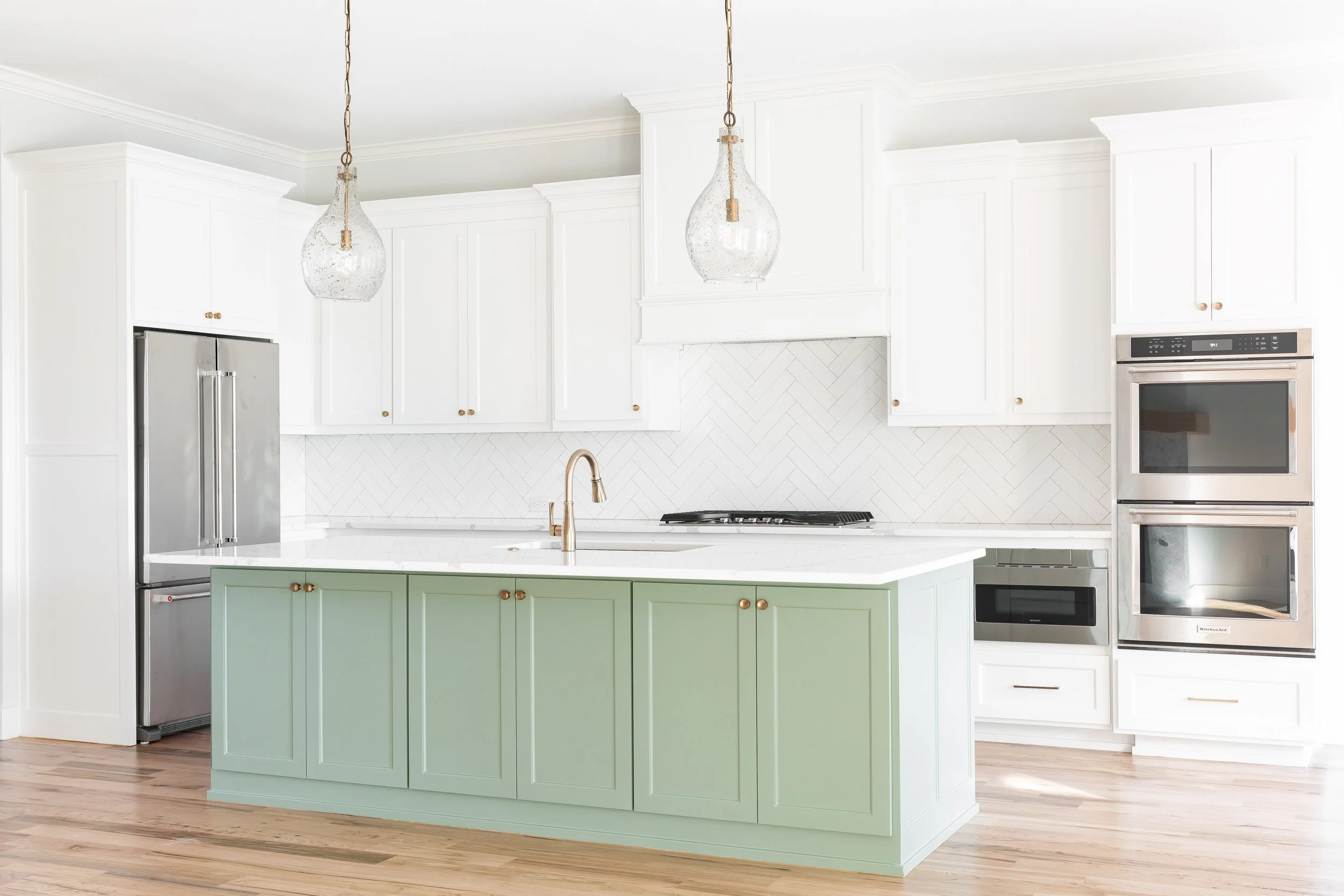 Modern white kitchen with a light green island, stainless steel refrigerator, wall oven, built-in microwave, and pendant lights.