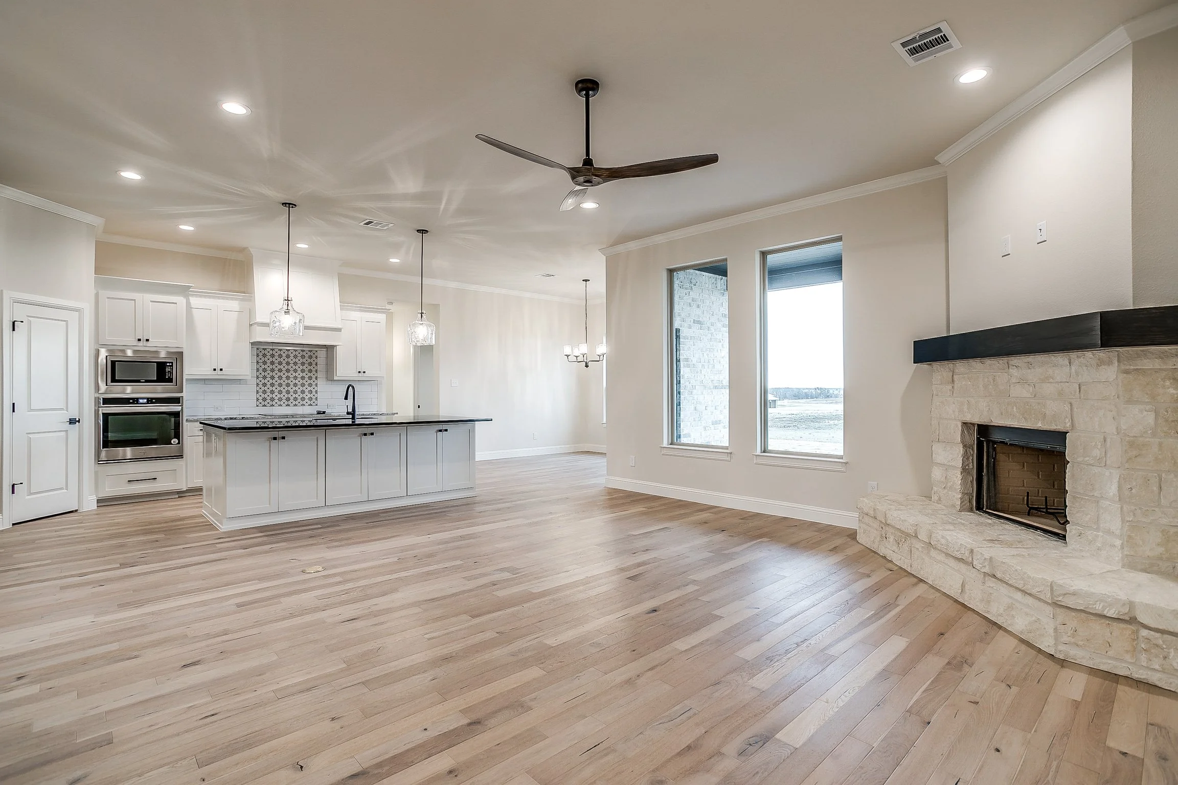 Open-concept kitchen and living area with light wood flooring, white cabinets, stainless steel appliances, a stone fireplace, and large windows letting in natural light.