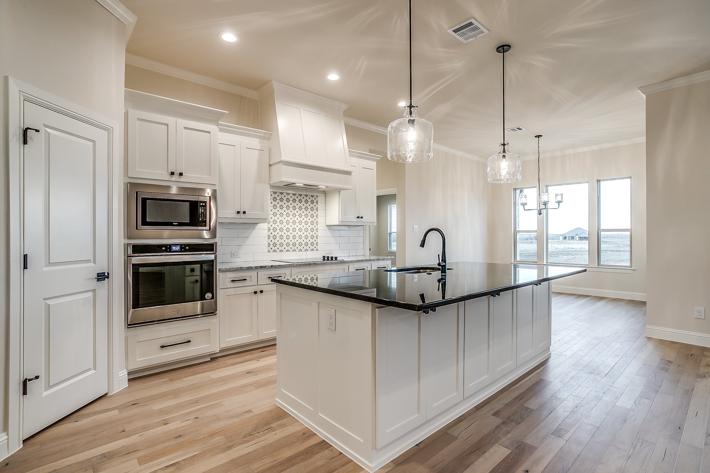Open-concept kitchen with white cabinets, a black countertop island, stainless steel microwave and oven, pendant lights, large windows, and hardwood floors.