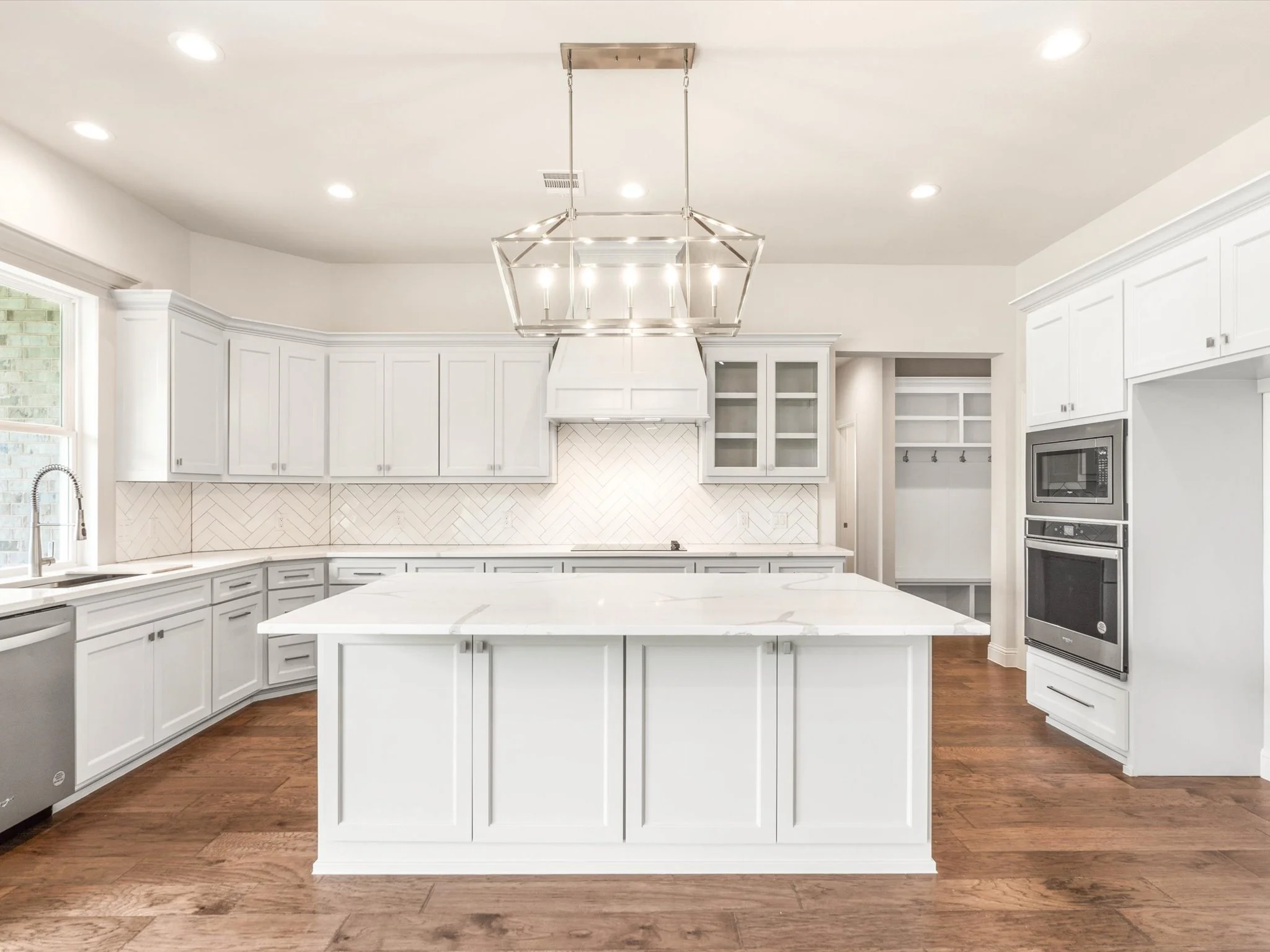 Modern kitchen with white cabinets, a white island, stainless steel appliances, and a geometric white tile backsplash. Recessed lighting and a large chandelier hang above the island. Hardwood floors.