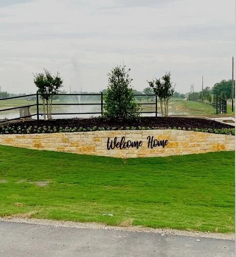 A landscaped yard with a stone wall that has a sign reading 'Welcome Home.' There are three trees behind the wall, a black metal fence, and a body of water in the background.