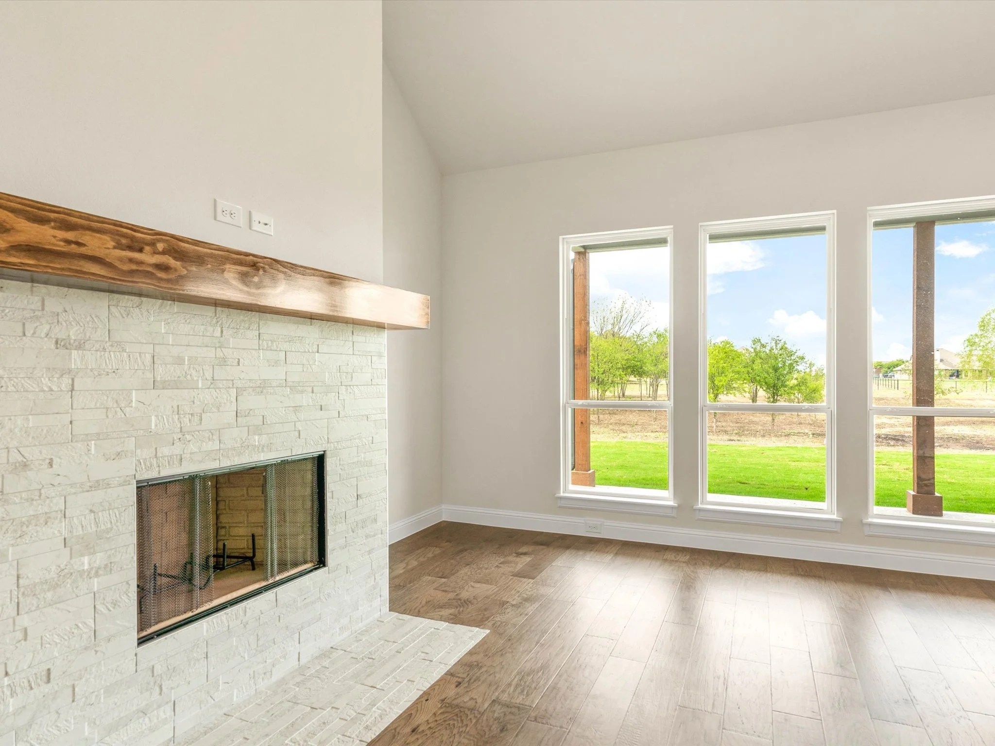 Interior of a modern living space with white walls, a stone fireplace with a wooden mantel, large windows showing a green outdoor landscape, and hardwood flooring.