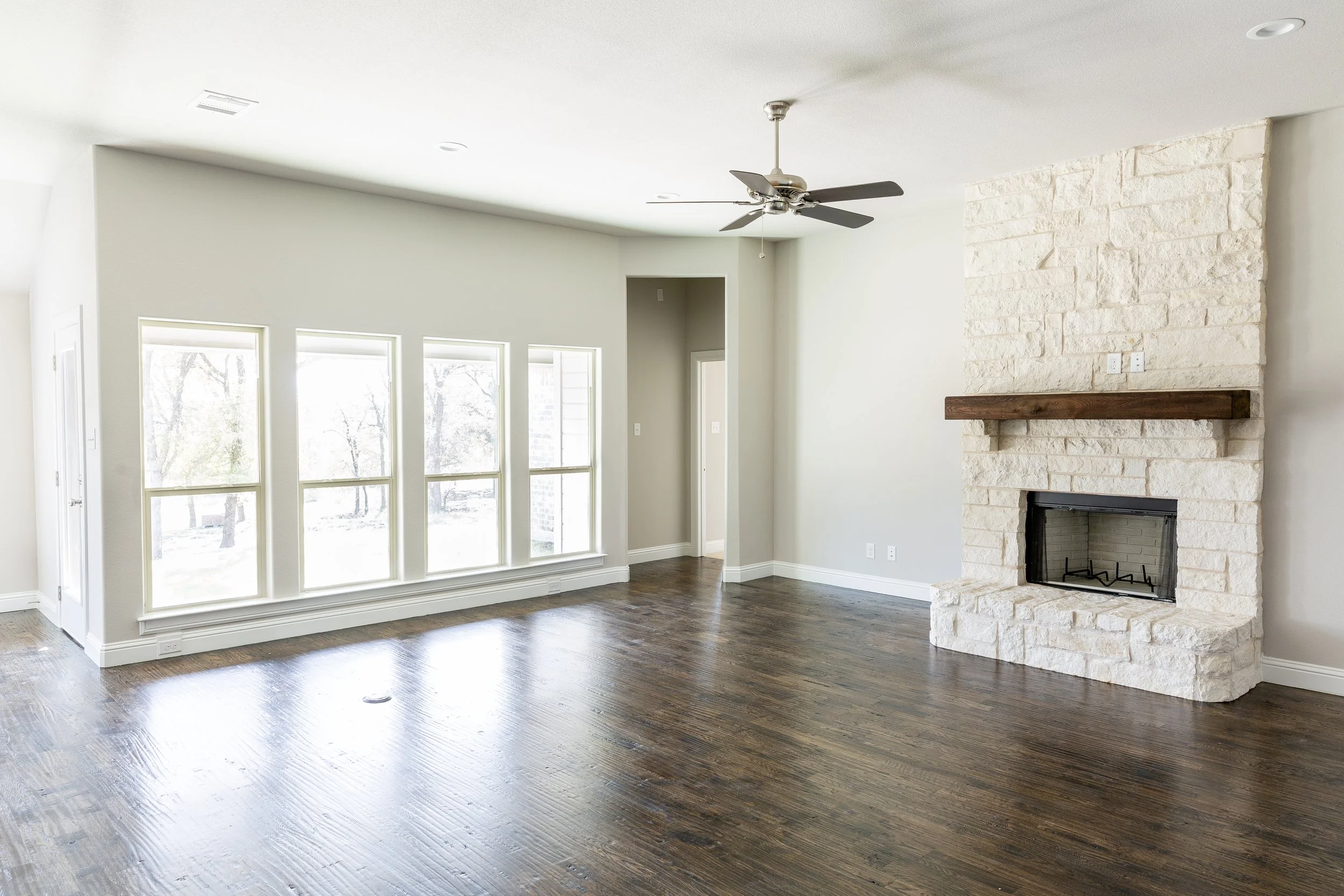 Empty living room with large windows, a stone fireplace with wooden mantel, ceiling fan, and hardwood floors.