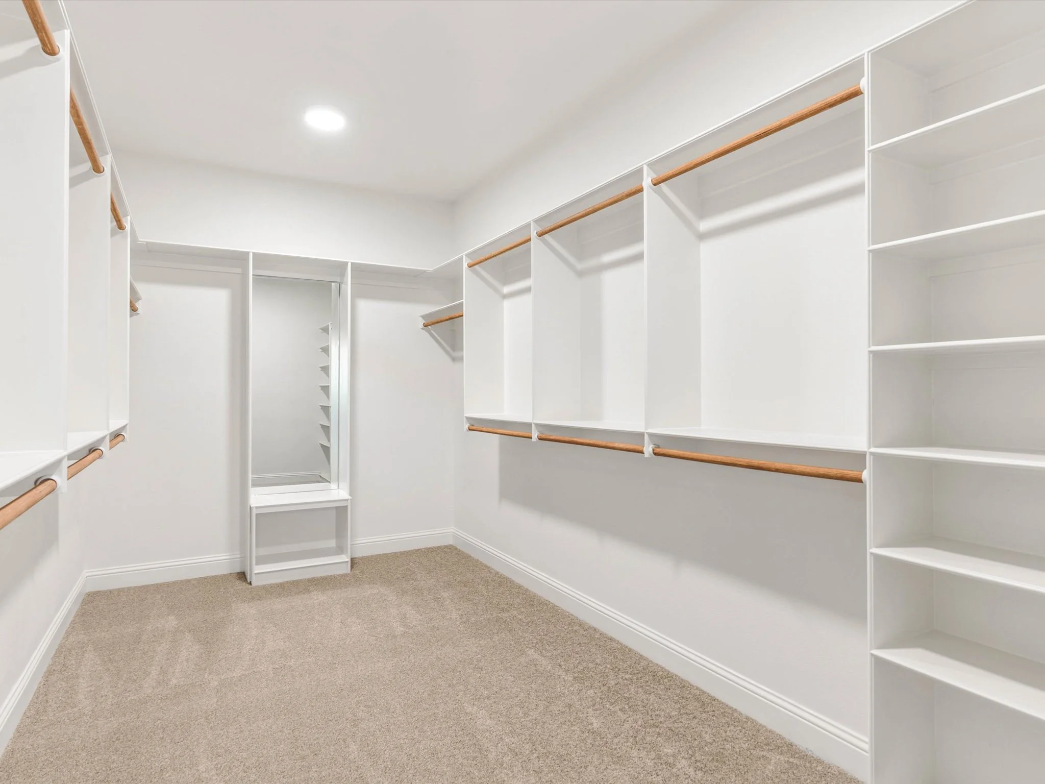 Empty walk-in closet with white shelving, wooden rods for hanging clothes, carpeted floor, and a mirror at the back wall.