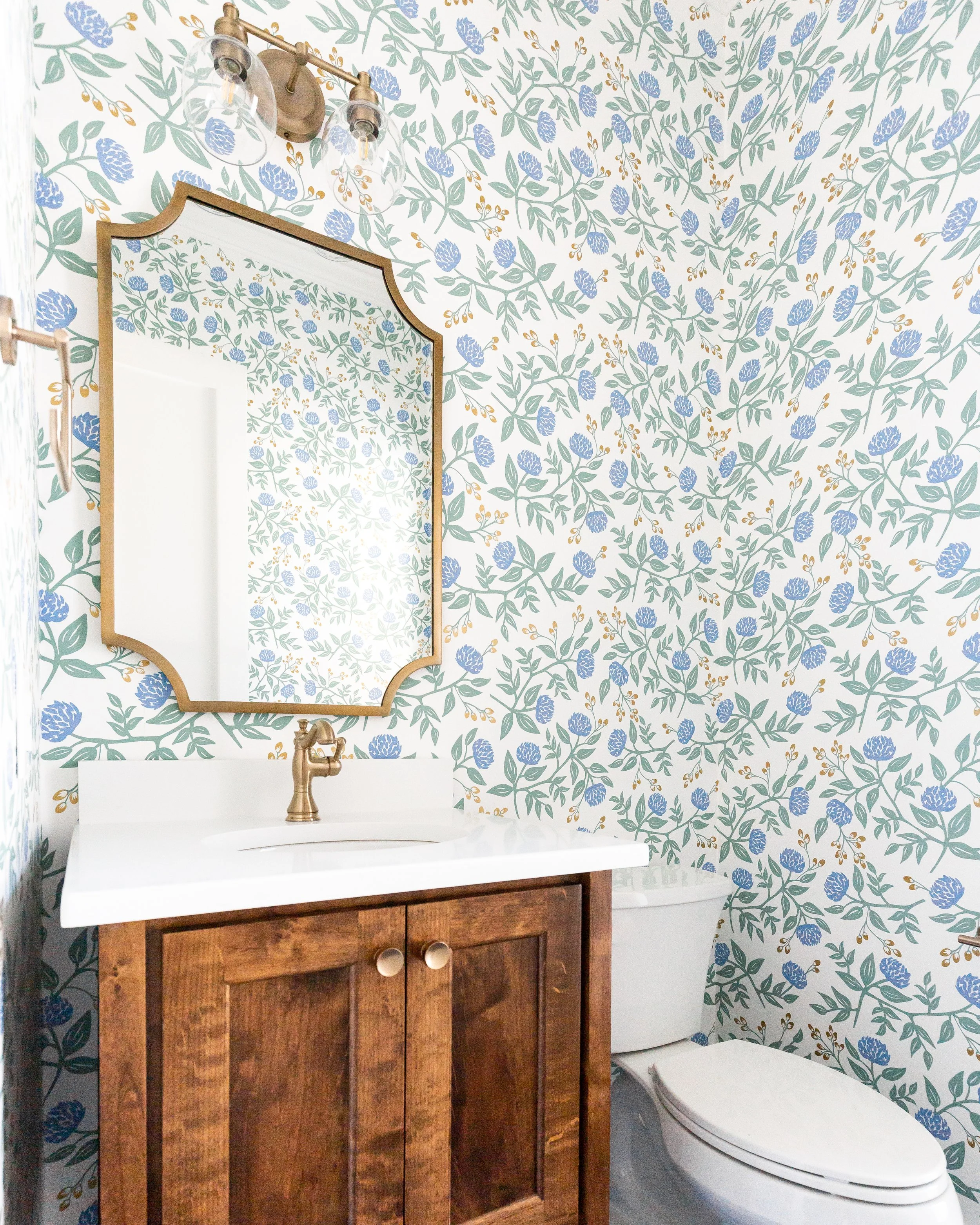 Bathroom with wallpaper featuring blue flowers and green leaves. A white vanity with a mounted gold faucet. A mirror with a gold frame above the vanity. A wall-mounted light fixture with three bulbs and glass shades. Toilet to the right of the vanity