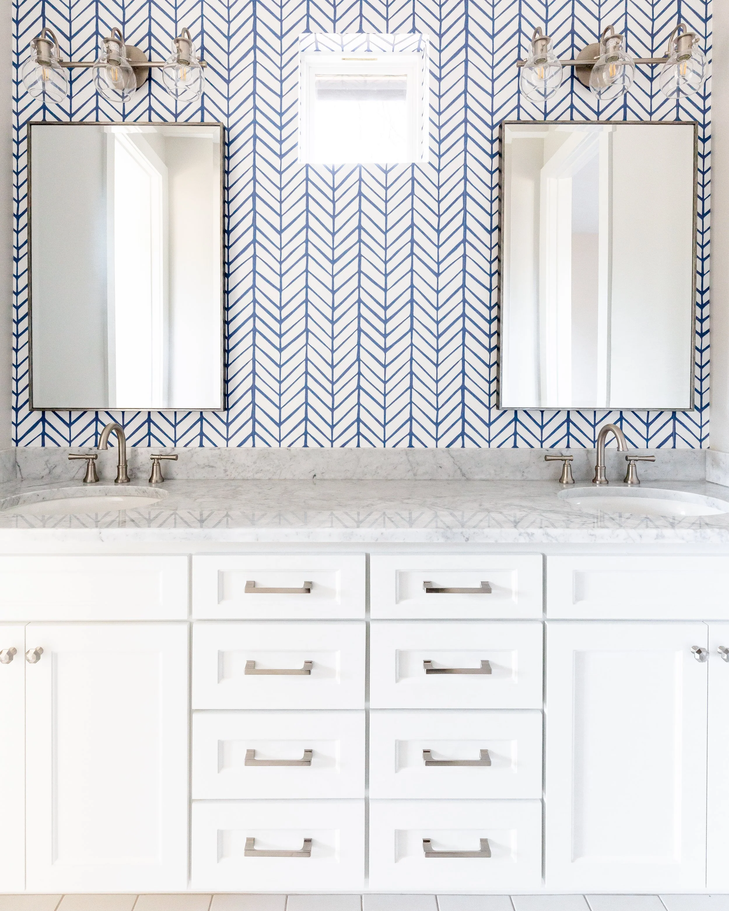Bathroom vanity with a marble countertop, two sinks, and a white cabinet with multiple drawers. Two mirrors are mounted above the sinks, and a blue and white patterned wall is behind them. A small square window is centered above the mirrors, with a w