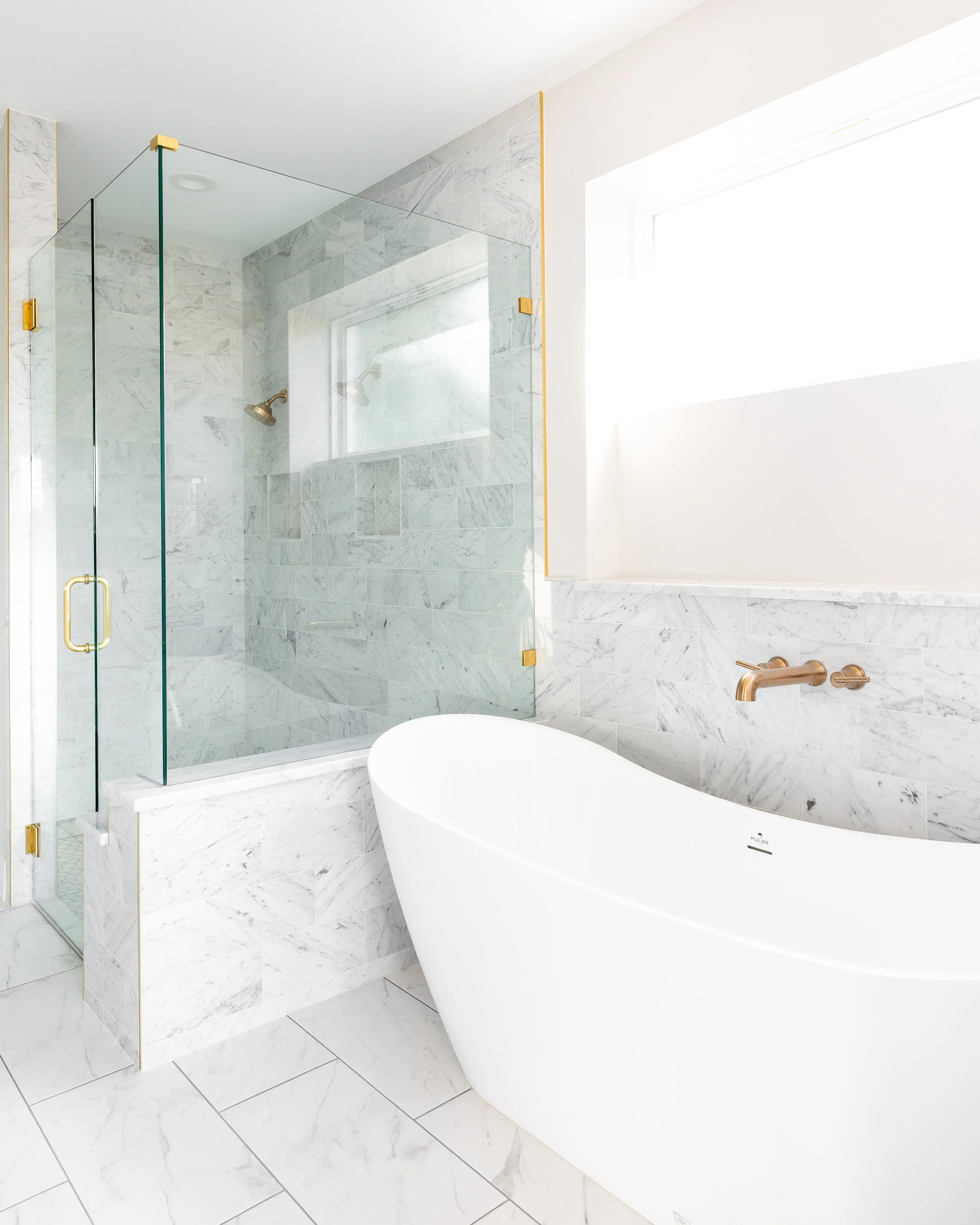 Modern bathroom with a white free-standing bathtub, marble tiled walls, a glass shower enclosure with brass hardware, and a large window allowing natural light.