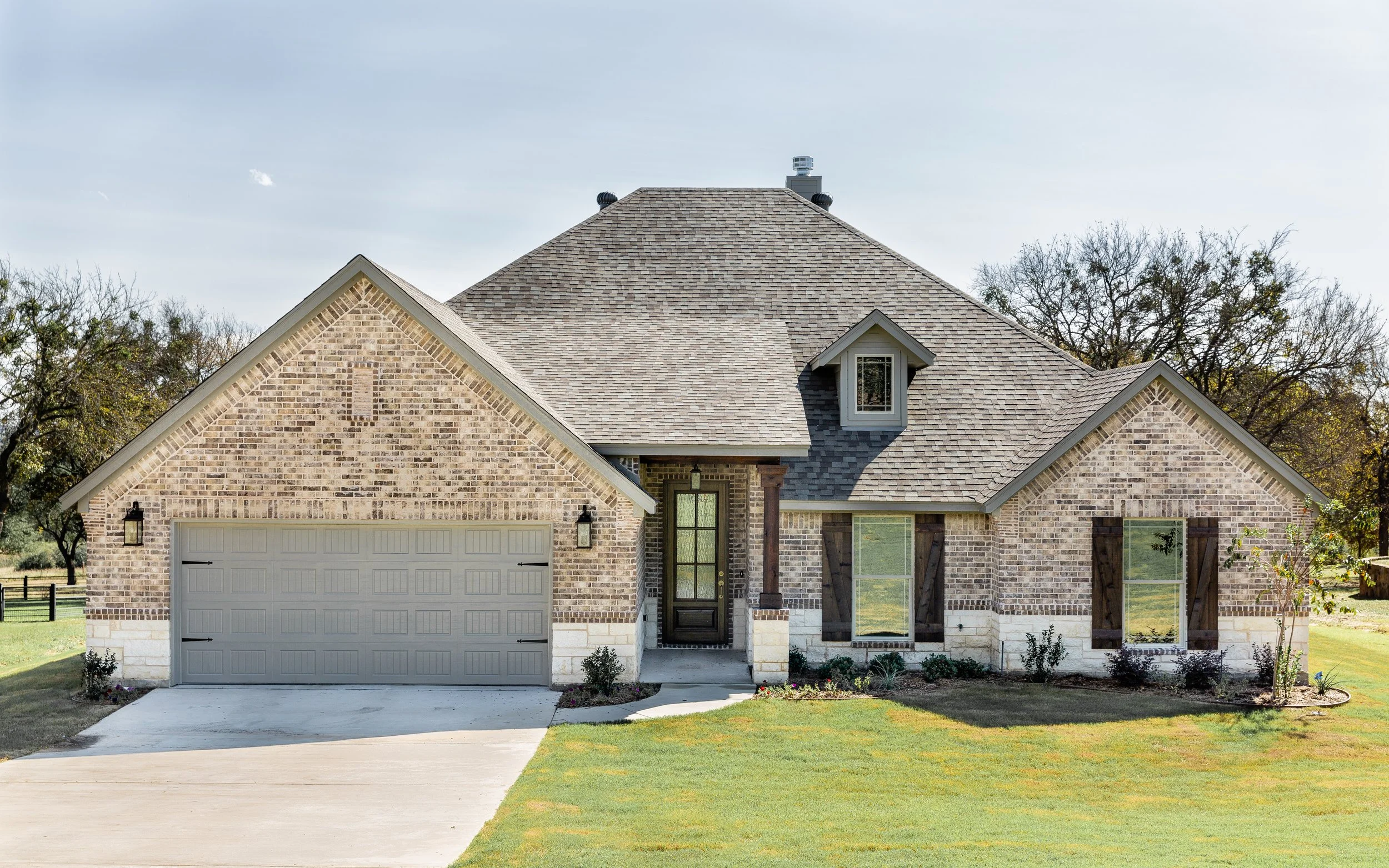 Front view of a two-story brick house with a gabled roof, attached garage, front door with a window, dormer window, and a front yard with grass and small bushes.