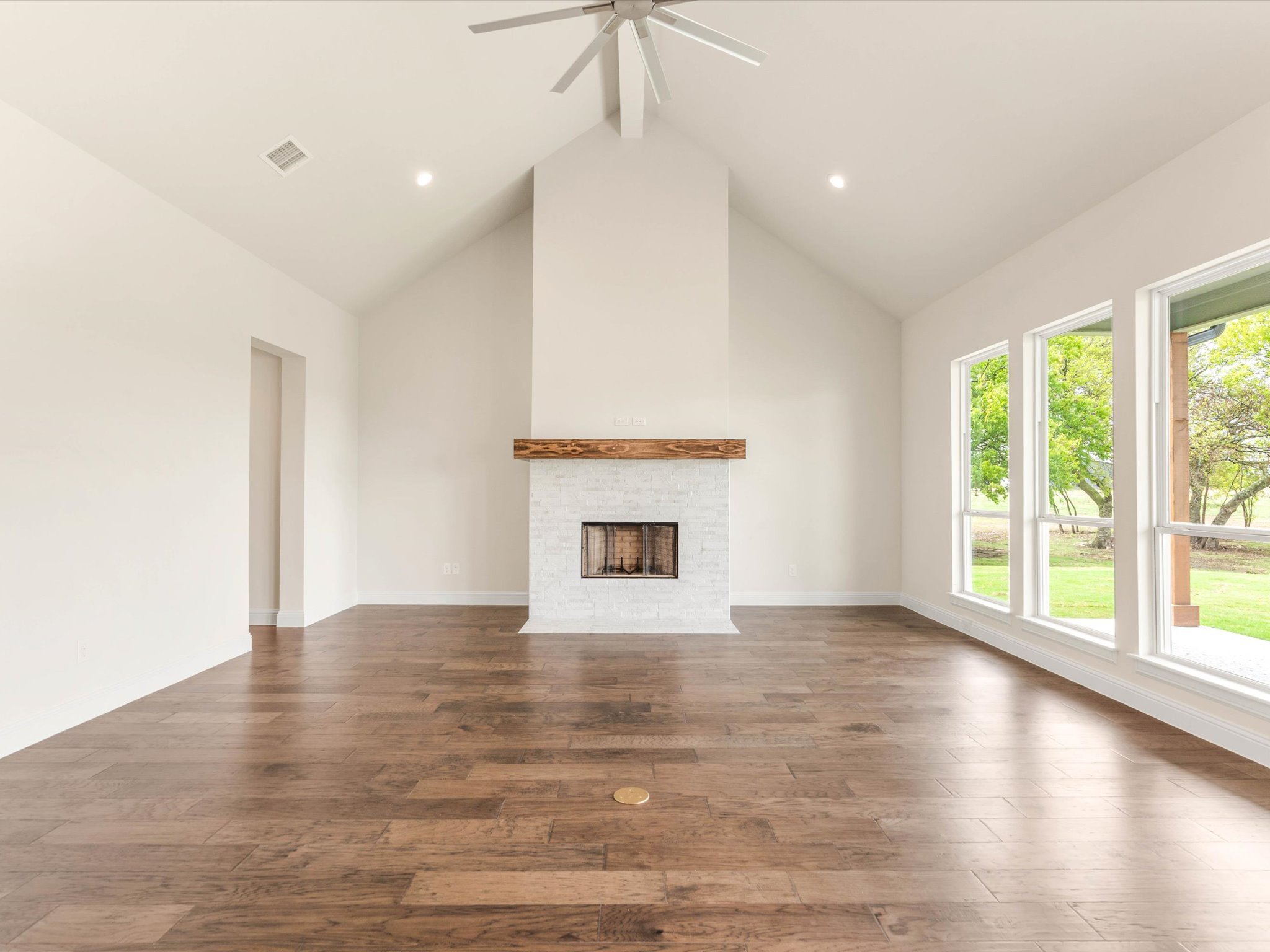 Empty living room with white walls, large windows showing trees outside, a white brick fireplace with a wooden mantel, and hardwood floors.