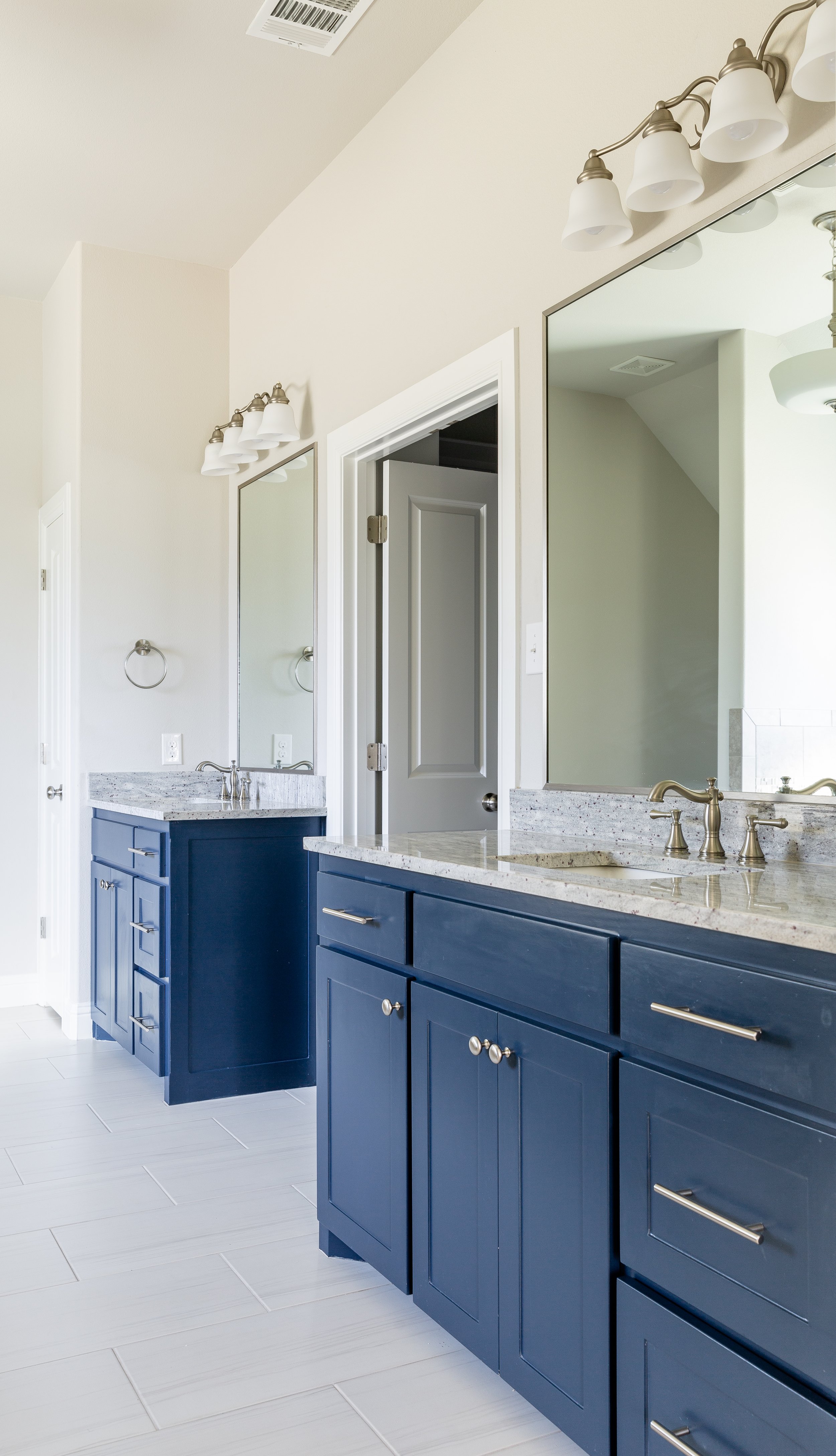Bathroom with navy blue cabinets, marble countertops, large mirror, white tile flooring, and wall-mounted light fixtures.