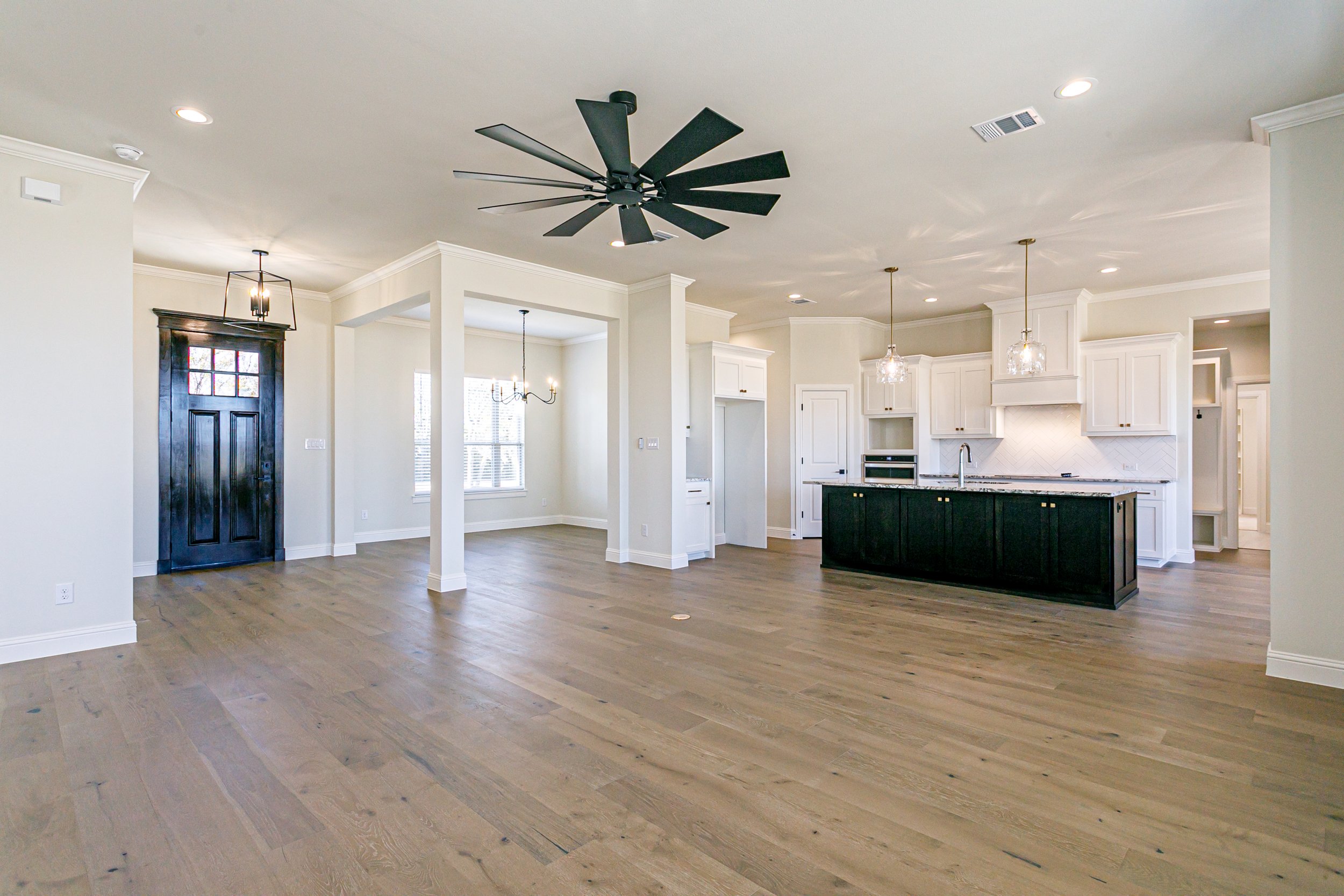 A spacious, open-plan kitchen and living area with hardwood floors, white walls, a black ceiling fan, and modern lighting fixtures. There is a black front door with a grid of glass panes, and large windows allowing natural light to fill the space.