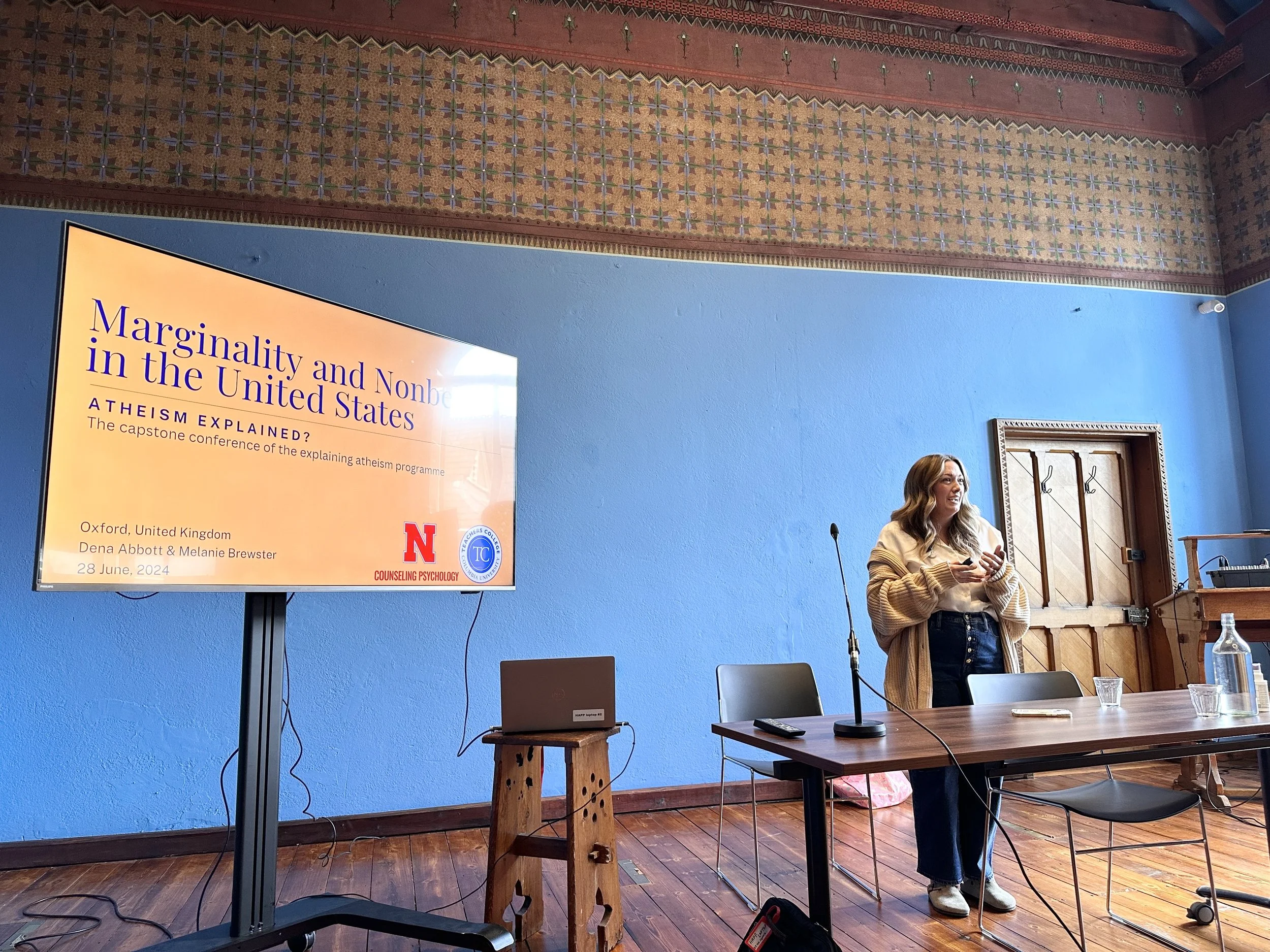 Dr. Abbott, a woman with long brown hair wearing a beige jacket is speaking at a podium during a presentation in a small blue room at the Oxford Museum of Natural History. The slide says 'Marginality and Nonbelief in the United States'.