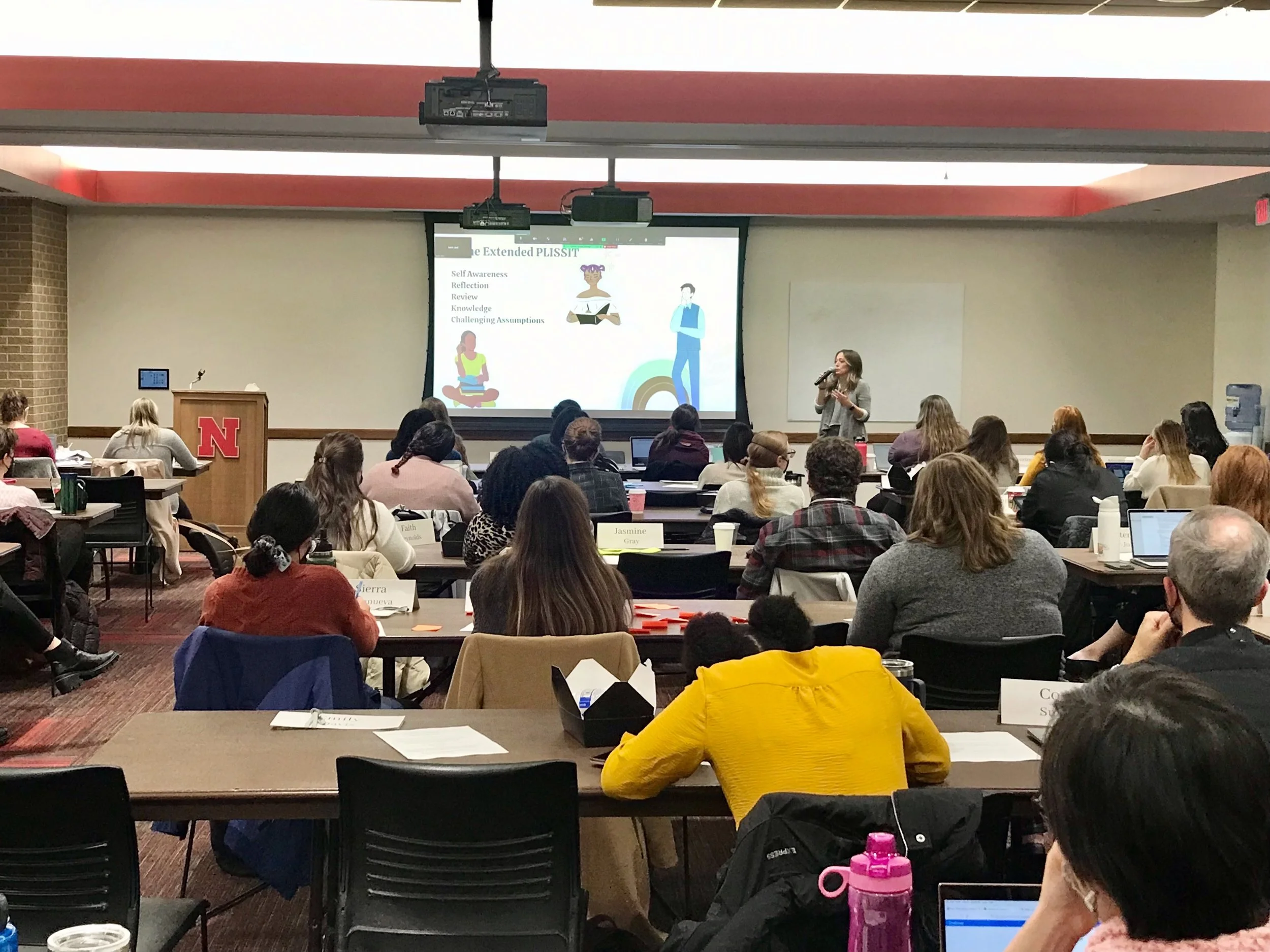 A large conference room filled with people attending a presentation by Dr. Abbott, a woman who is standing on the right speaking into a microphone. There is a large screen behind her displaying presentation slides titled 'The Extended PLISSIT.'