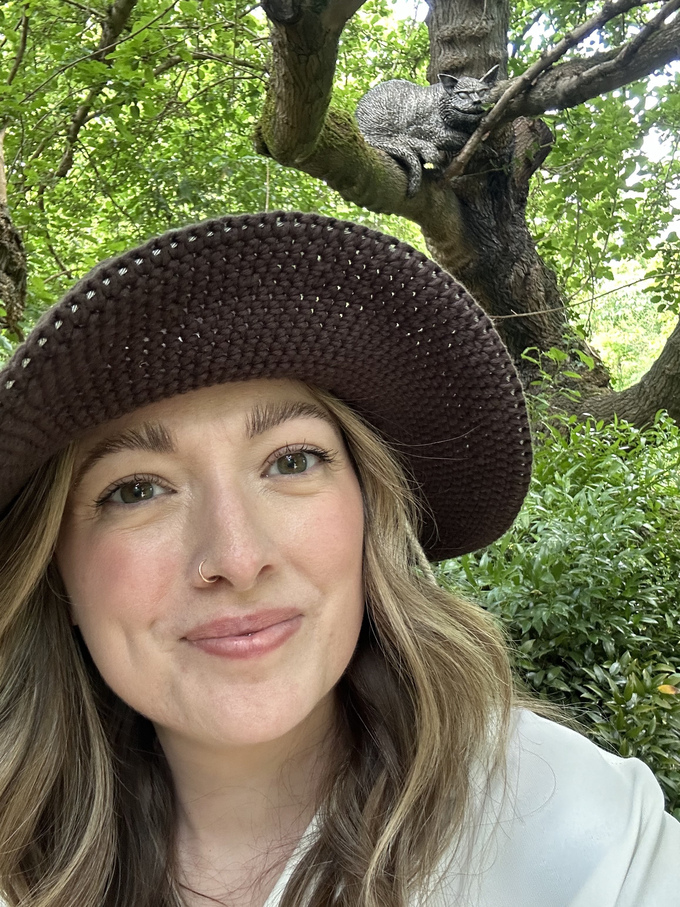Dr. Abbott, a woman with long blonde hair and a nose ring is smiling at the camera. She is wearing a wide-brimmed straw hat. Behind her, there are green leaves and branches of a tree, with a gray cat perched on a branch.