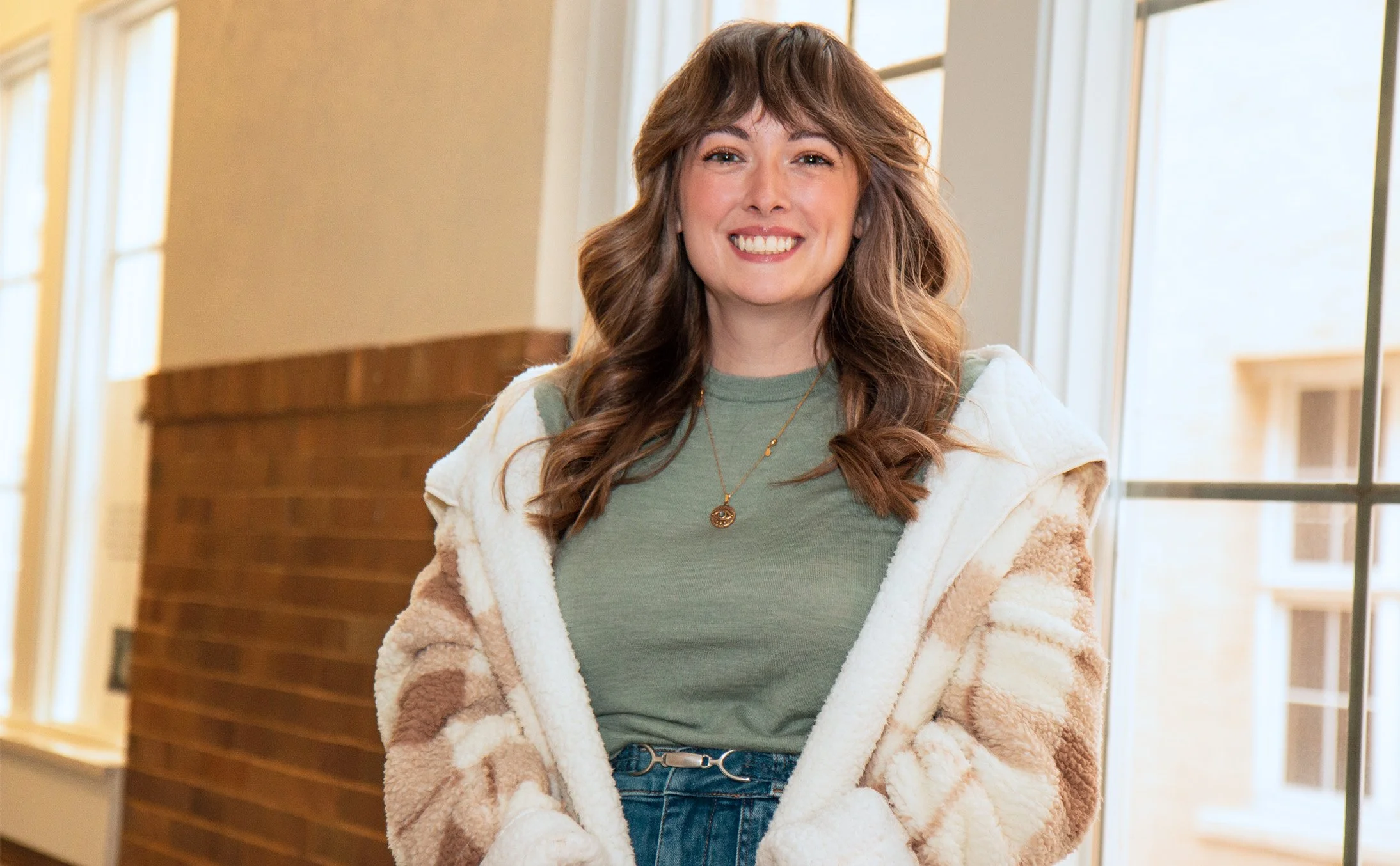 Dr. Abbott, a woman with long, wavy brown hair wearing a green shirt, layered with a cream and beige patterned fleece jacket, standing indoors near a window with sunlight streaming in.