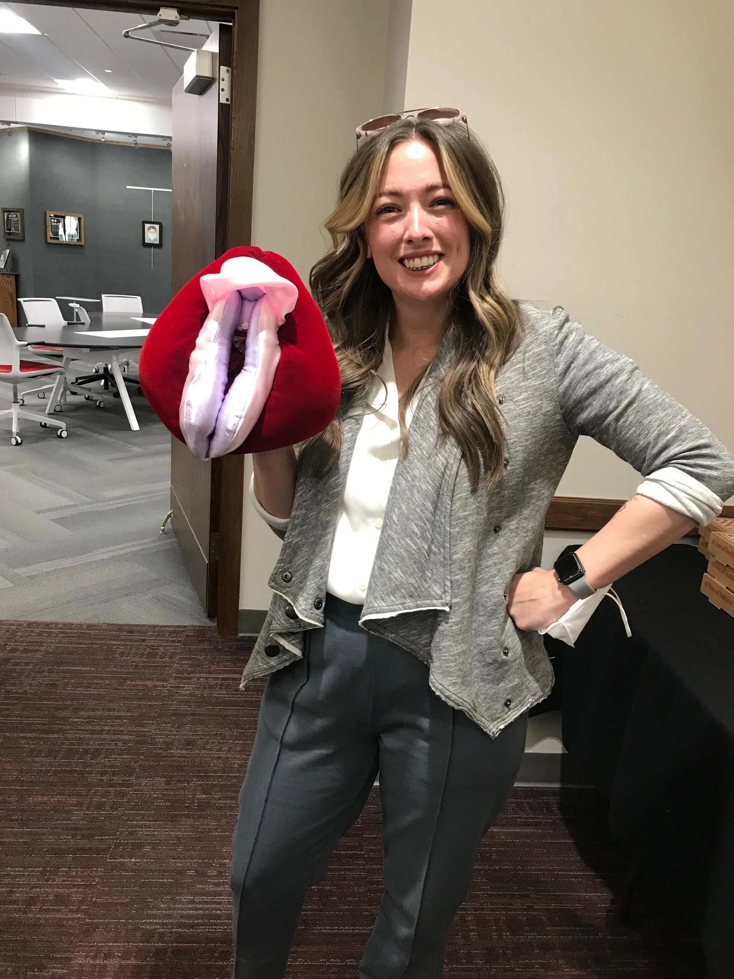 Dr. Abbott, a woman smiling, holding a stuffed vulva used for sex ed demonstrations. She has long, wavy hair and is wearing a gray blazer over a white shirt and gray pants.