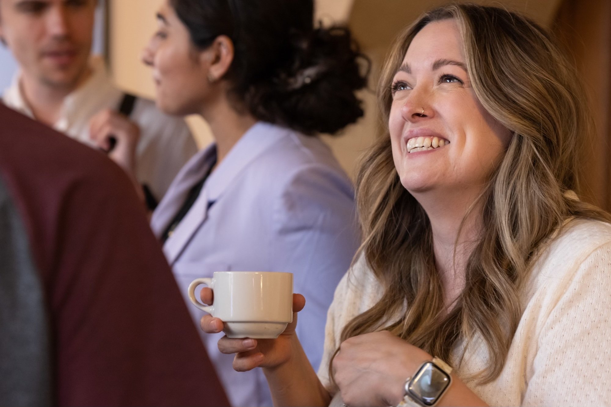Dr. Abbott, a woman with long wavy hair and a nose ring smiling while holding a white coffee mug, engaged in conversation with a person; other people are in the background.