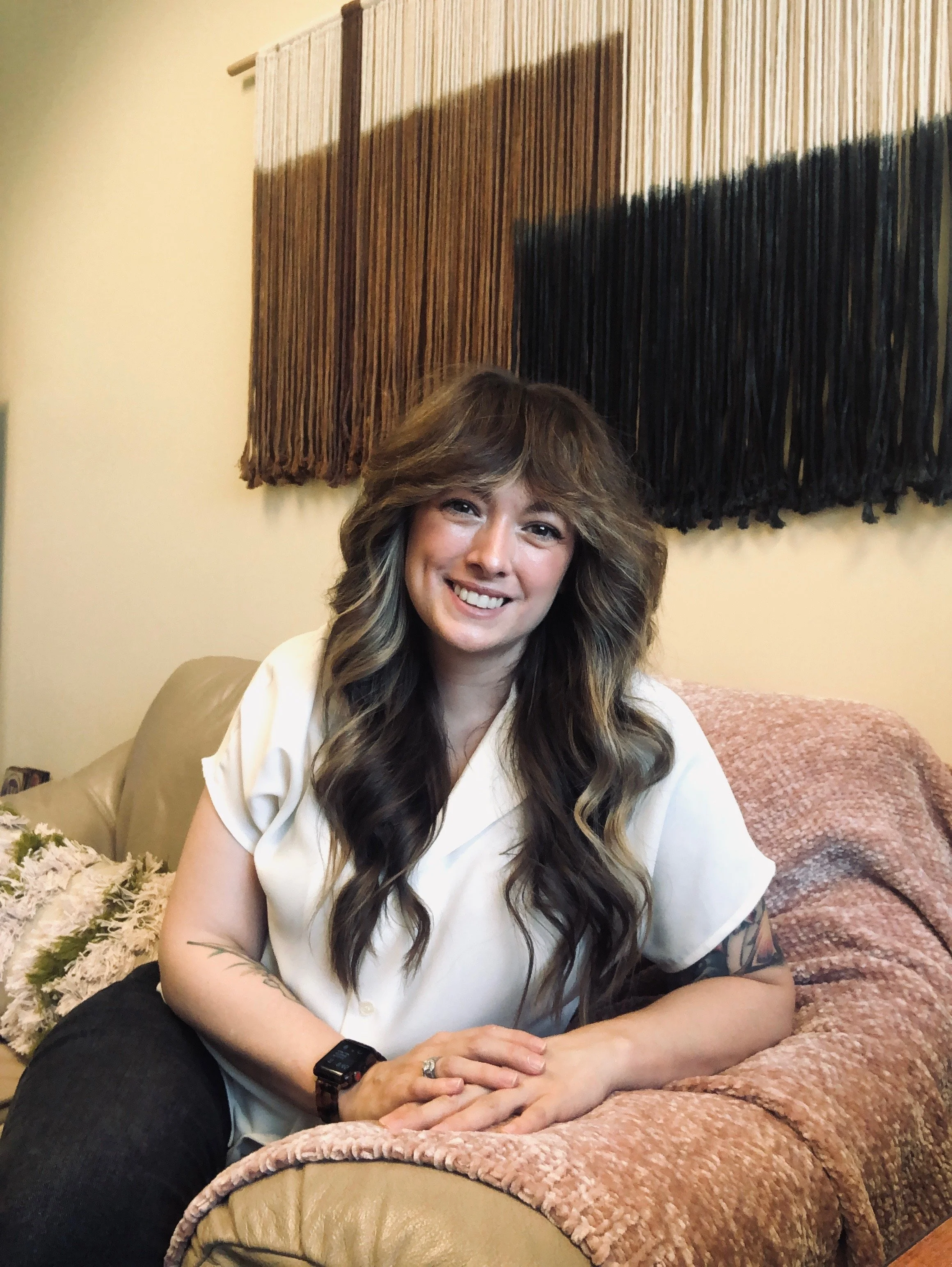 Dr. Abbott, a woman with long wavy hair sitting on a beige couch. Behind her, there is a peach-colored blanket and a decorative wall hanging with vertical brown and black strips.