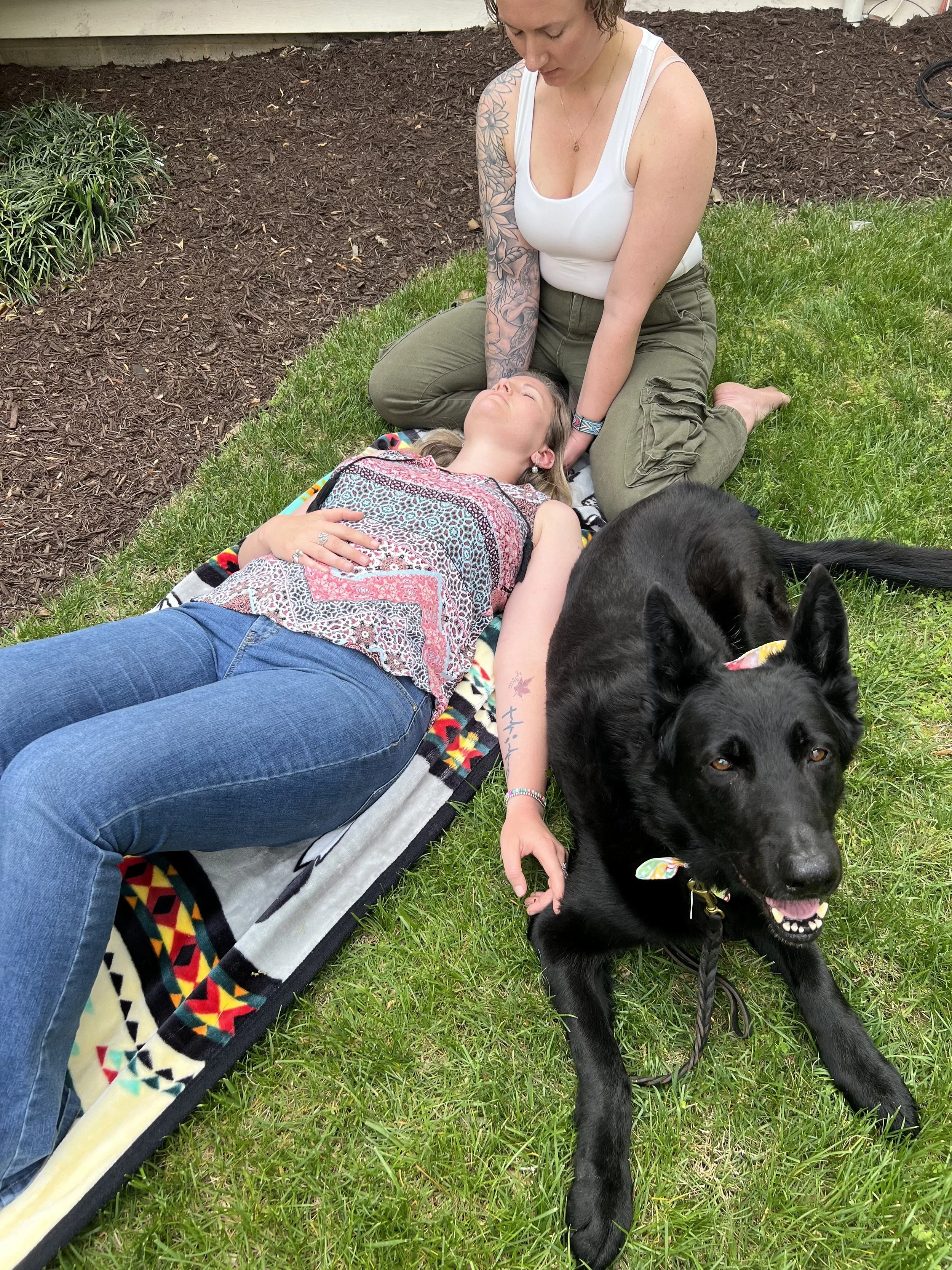 A woman lying on the grass with a black dog and another woman kneeling nearby, petting the dog, in a backyard with mulch and plants.
