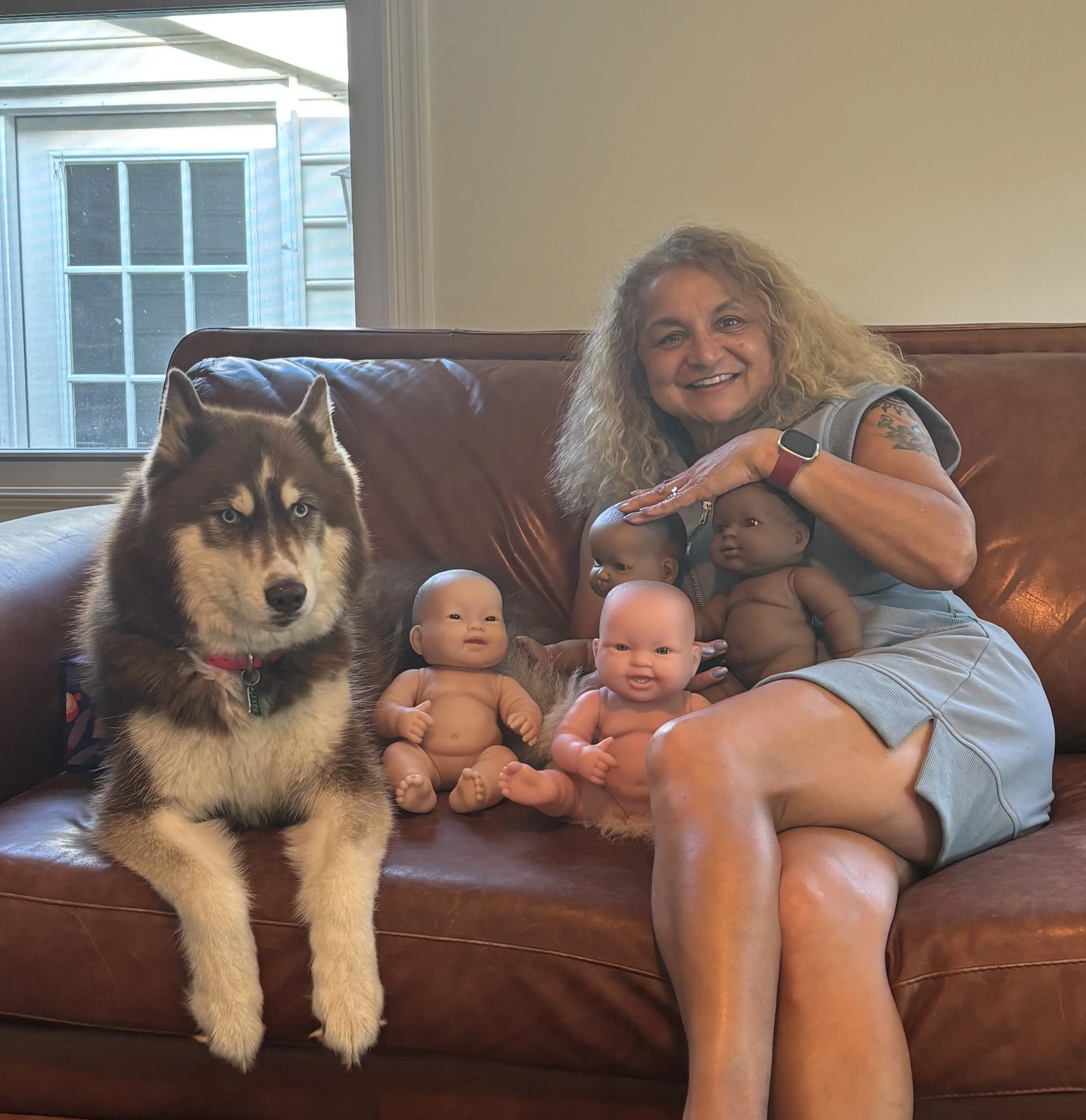 A woman smiling on a brown leather couch, with a Siberian Husky dog and three dolls, inside a room with a window showing an exterior view of a house.
