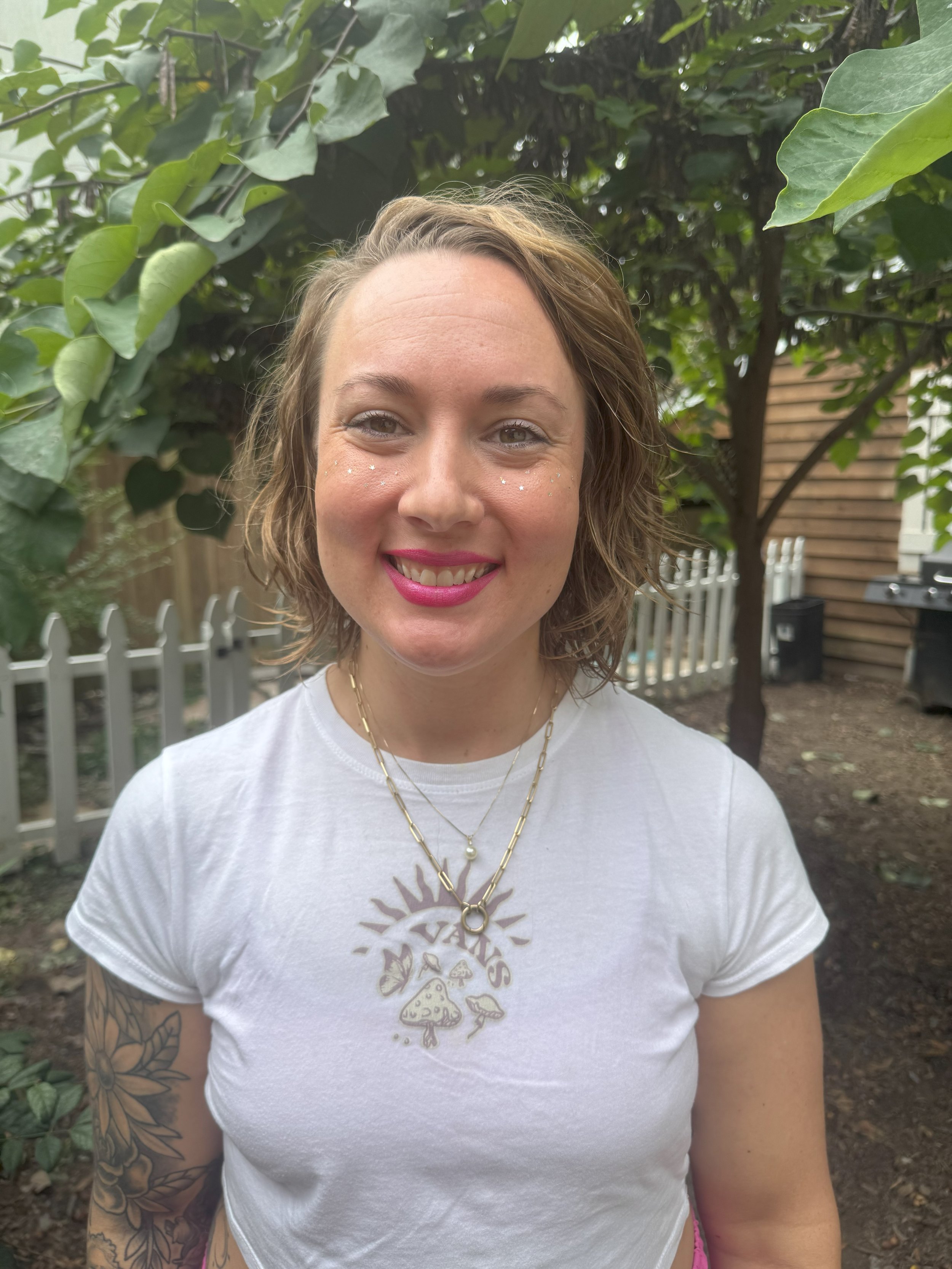 A woman with wavy light brown hair and pink lipstick smiling outdoors in front of a tree and a wooden fence, wearing a white t-shirt with mushroom and sunflower graphic, layered gold necklaces, and visible tattoos on her arm.