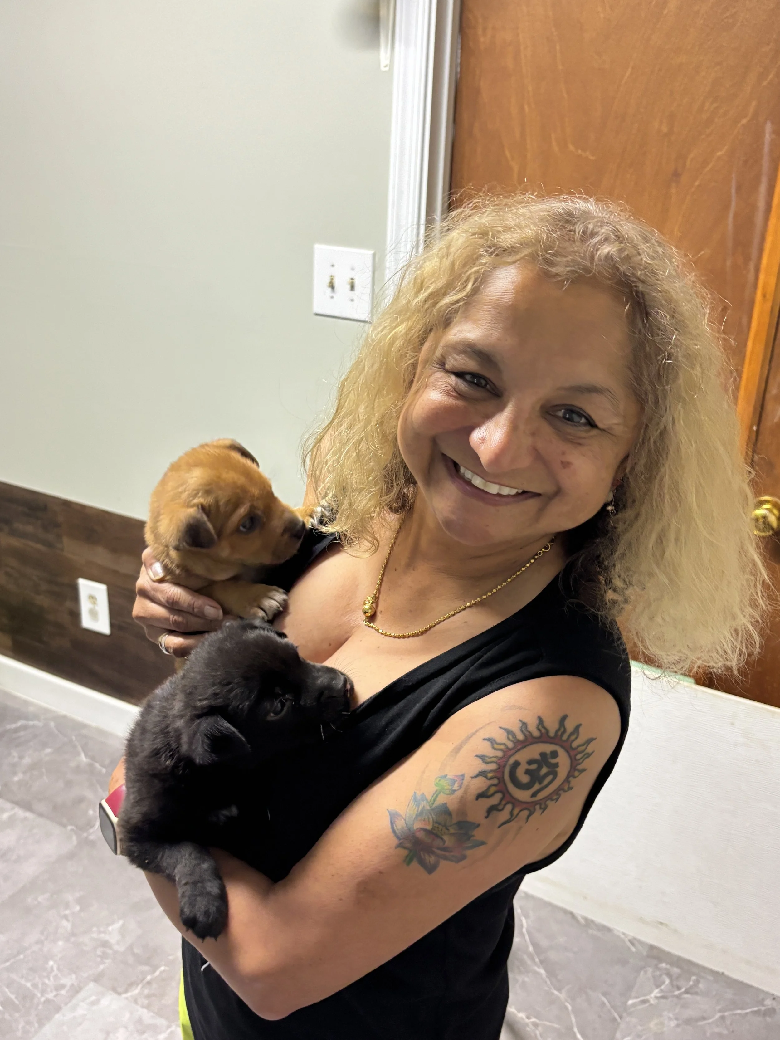 A woman with blonde curly hair smiling and holding three puppies, one black, one brown, and one dark brown, in an indoor setting with beige walls, wooden accents, and exposed electrical outlets.