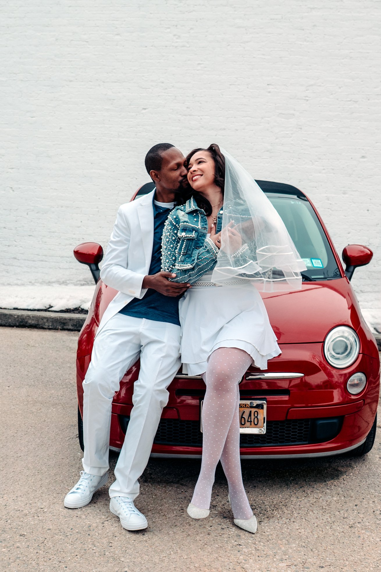 A couple in wedding attire sitting on the hood of a red car against a white brick wall. They are leaning in for a kiss, with the woman wearing a sheer veil, polka dot stockings, and a structured denim jacket. The man is wearing a white suit and white sneakers.
