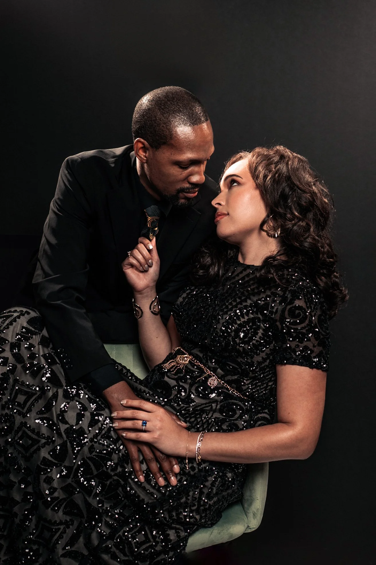A man and woman are close together, gazing into each other's eyes. The man is leaning over the woman, who is sitting on a light-colored chair, with a dark background. The woman is wearing a black, sparkly dress and jewelry, and the man is dressed in a black suit with a patterned tie.