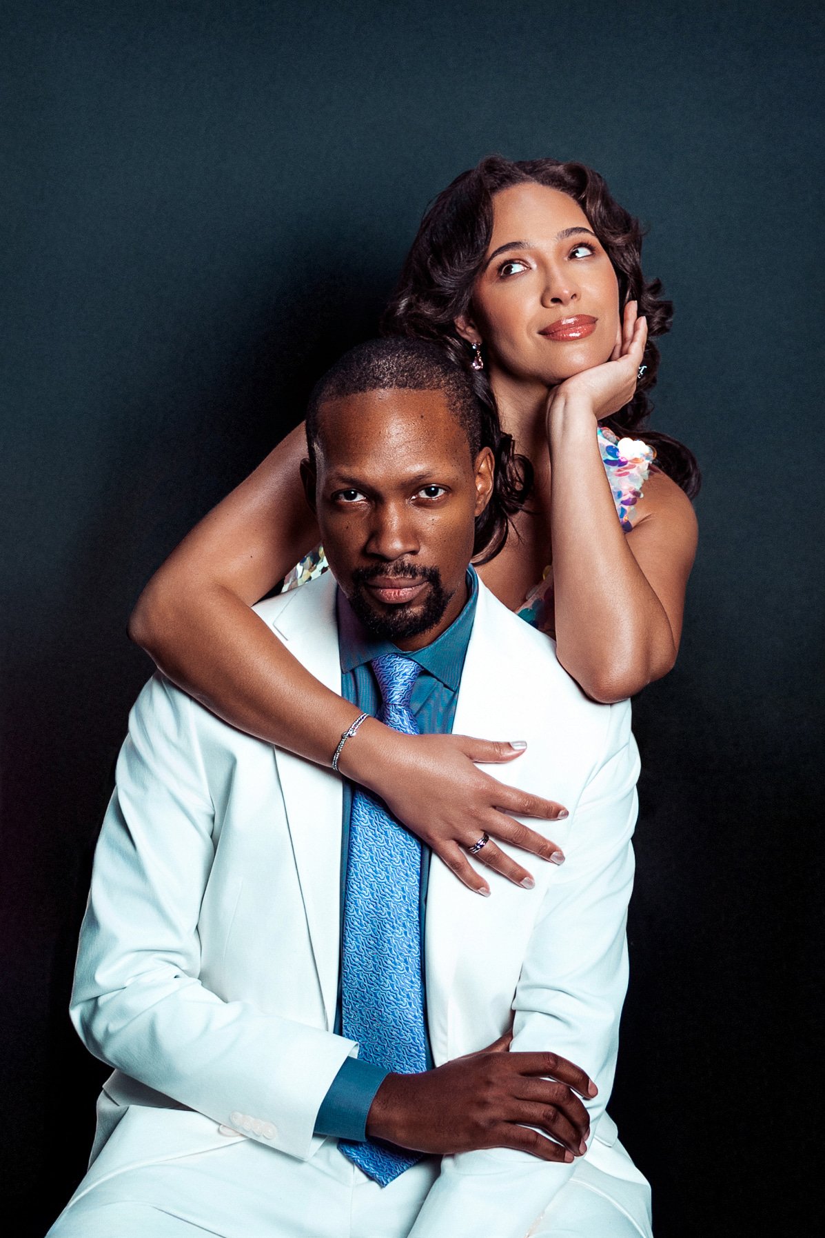 A man in a white suit with a blue tie seated, with a woman in a colorful dress resting her head and arm on his shoulder, both against a dark background.