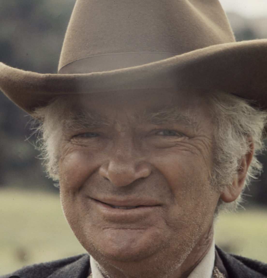 Close-up of Buddy Epsen with gray hair, wearing a cowboy hat, smiling outdoors.
